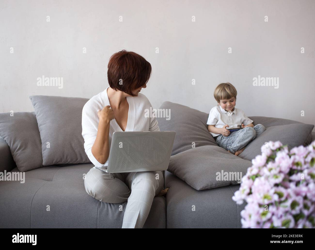 Mutter und Sohn mit digitalen Gadgets sitzen auf dem Sofa im Wohnzimmer. Unkenntliche Frau arbeitet zu Hause auf einem Laptop, neben einem Kind spielt Stockfoto