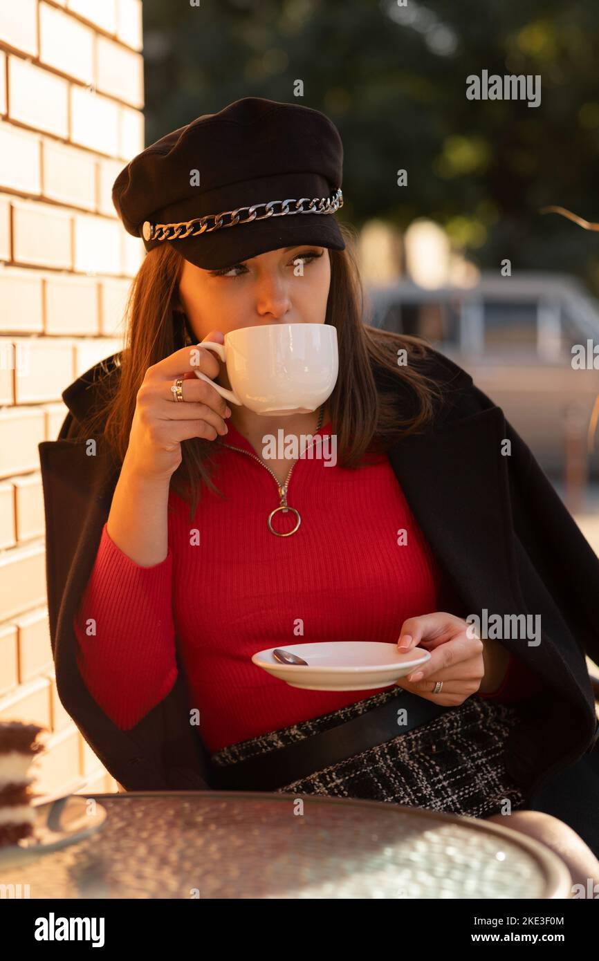 Nachdenkliches Mädchen in stilvollem schwarzen Hut und herbstlich leuchtendem roten Pullover, das in einem Pariser Café im Freien sitzt und Kaffee trinkt. Stockfoto