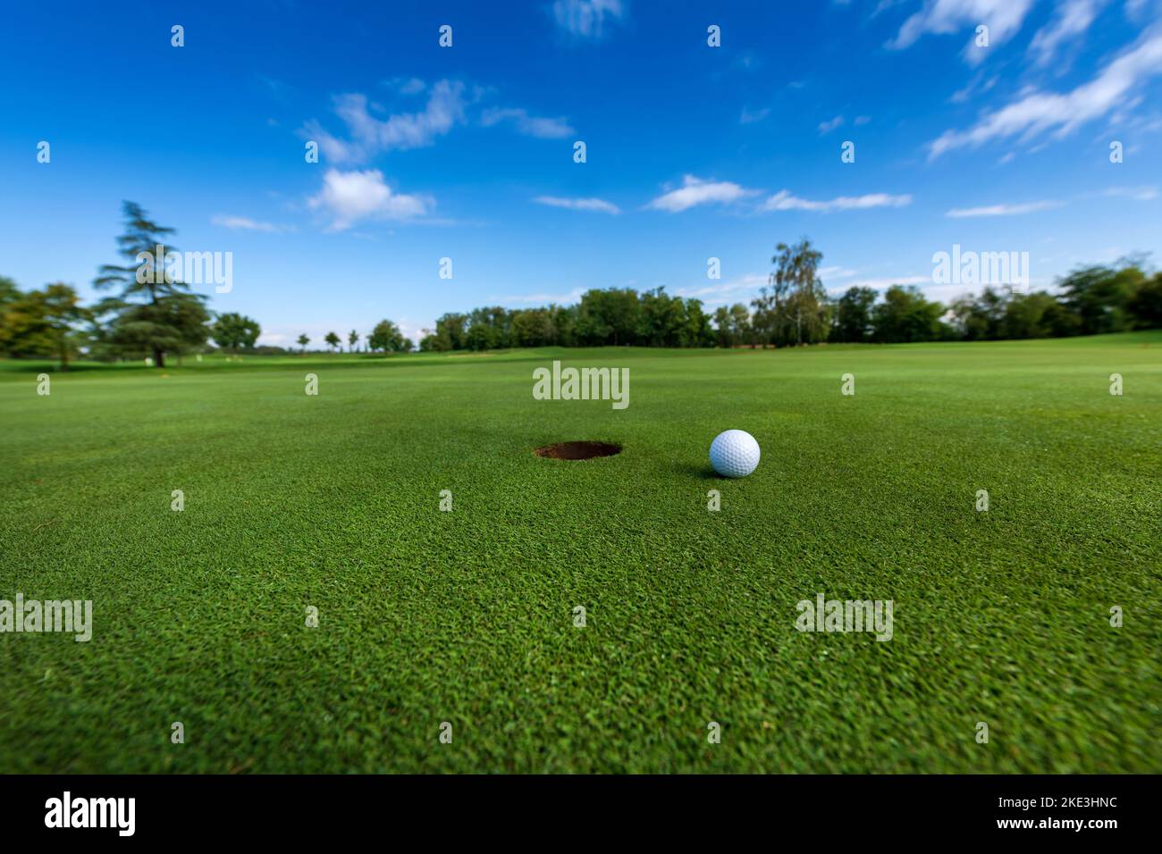 Weißer Ball auf grünem Gras in der Nähe von Loch im Boden gegen bewölkten blauen Himmel während des Golfspiels platziert Stockfoto