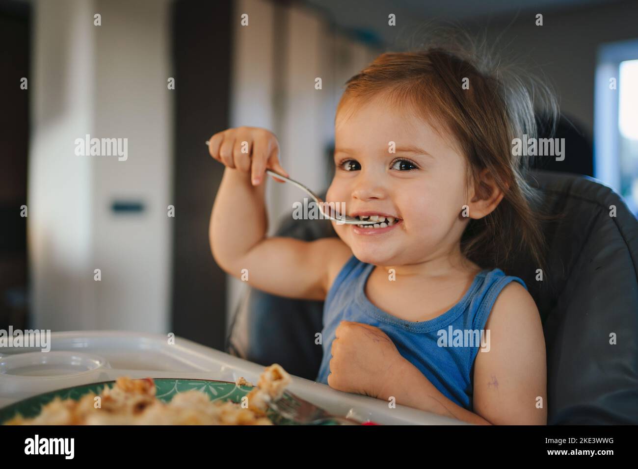 Baby sitzt im Hochstuhl mit Löffel und Teller in der Küche. Essen Sie selbst und machen Chaos. Gesunde Ernährung. Frohe Familie. Süßes Essen. Stockfoto