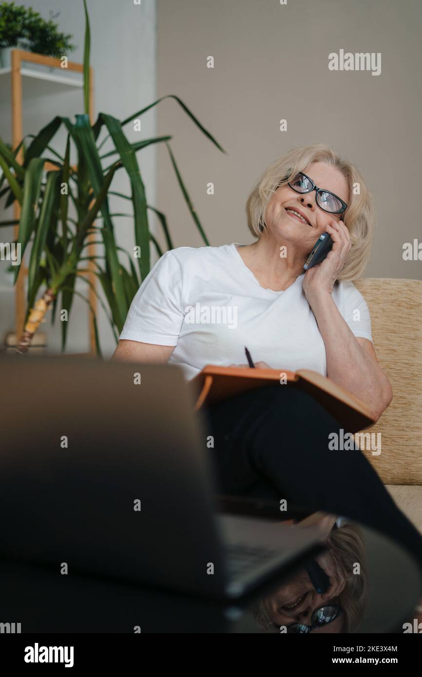 Moderne erfolgreiche ältere Frau mit Brille und Smartphone sitzt auf dem Sofa und spricht mit ihrer Freundin und arrangiert ein Abendessen. Internet Stockfoto