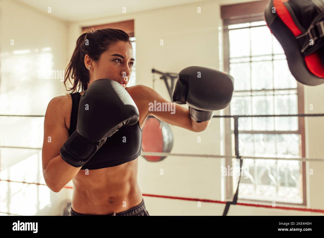 Boxerin stanzt Fokuspolster, während sie in einem Boxring steht. Sportliche junge Frau, die ein Boxtraining in einem Fitnessstudio absolviert. Stockfoto