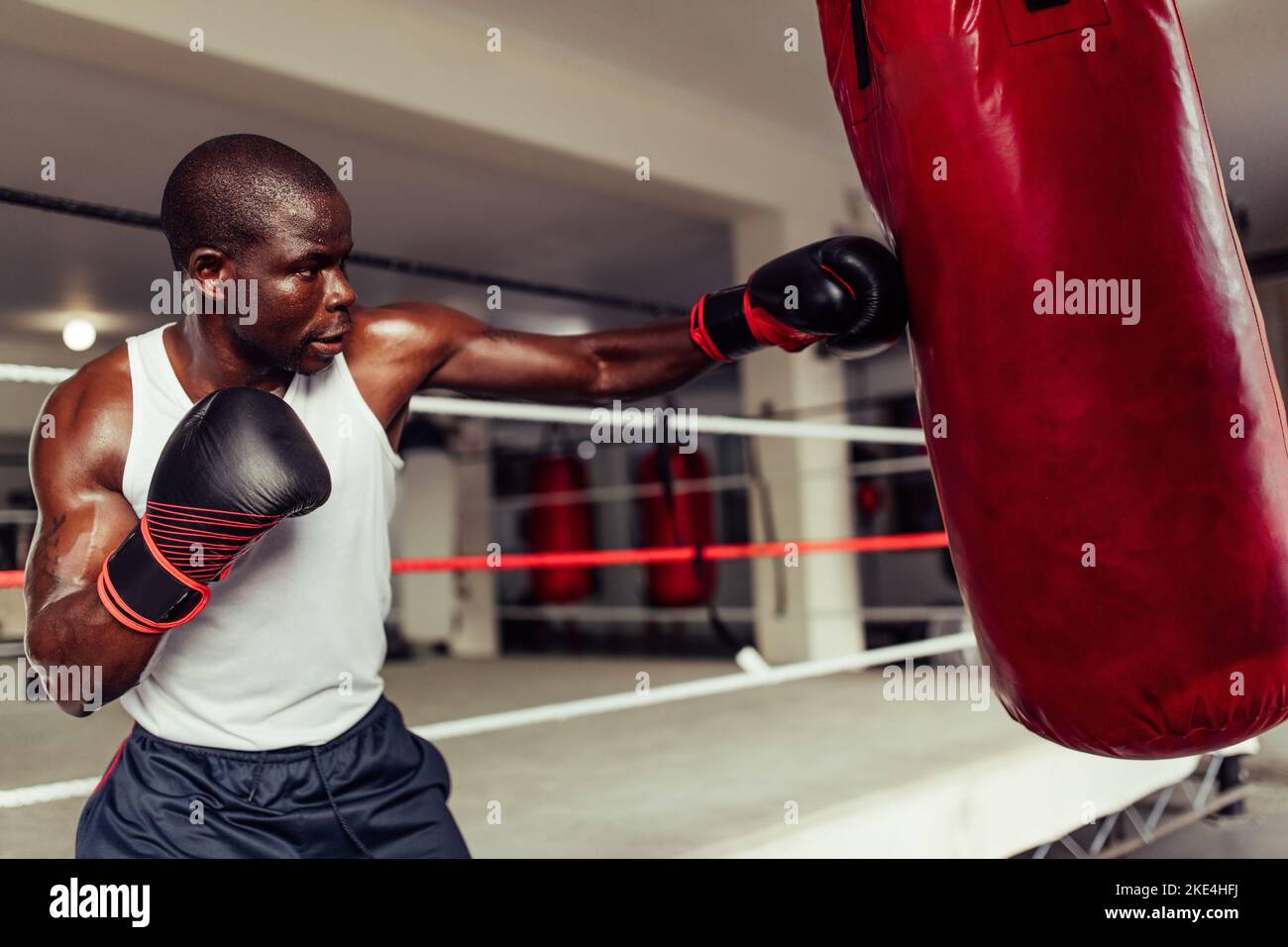 Fit afrikanischen Boxer werfen einen starken Schlag auf einen roten Leder Boxsack in einem Fitness-Studio. Sportlicher junger Mann, der in einer Boxhalle trainiert Stockfoto