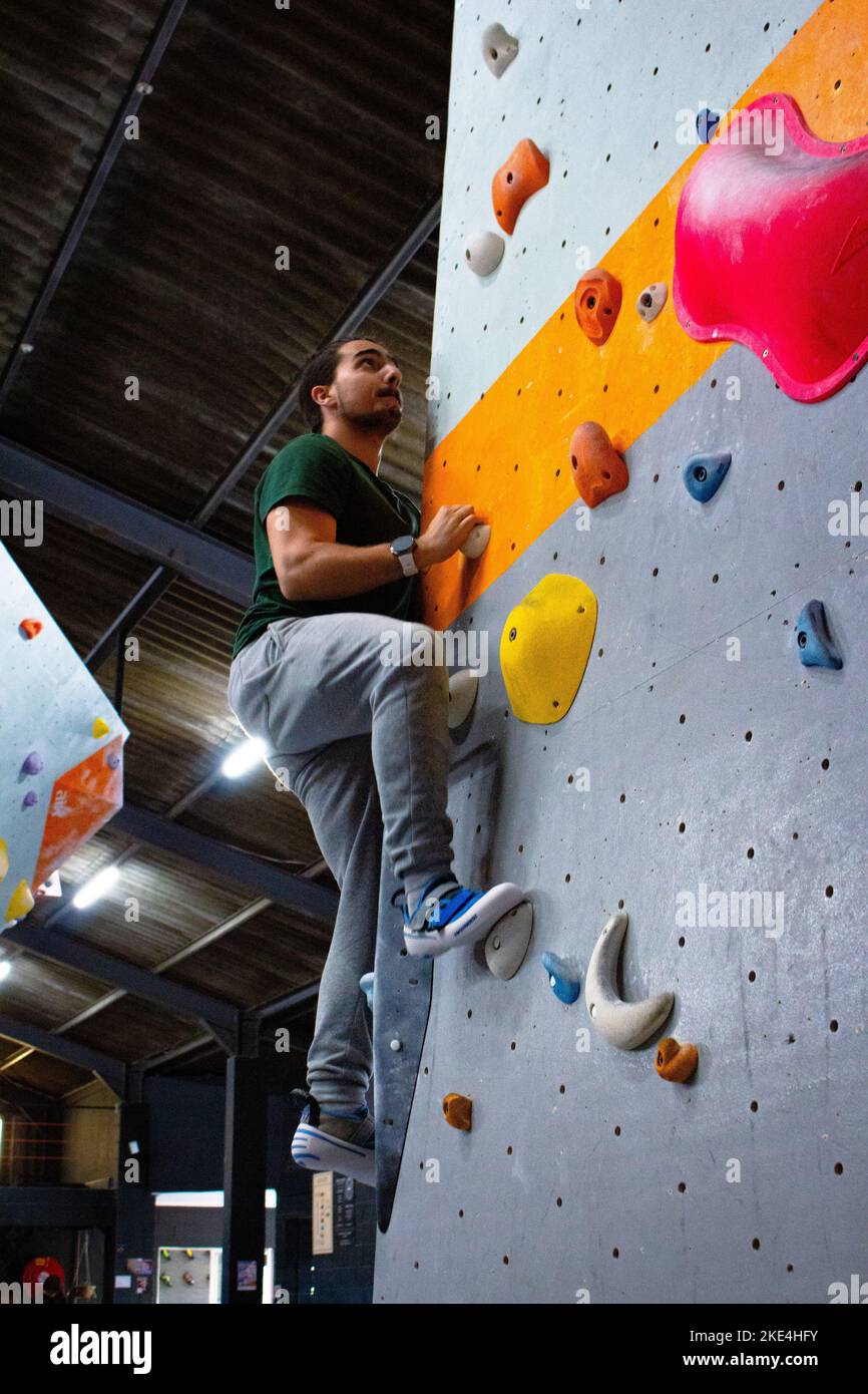 Ein hübscher Rock-Climber-Mann, der auf bunten Slopern aufsteigt, um die Kletterwand im Fitnessstudio zu bouldern Stockfoto