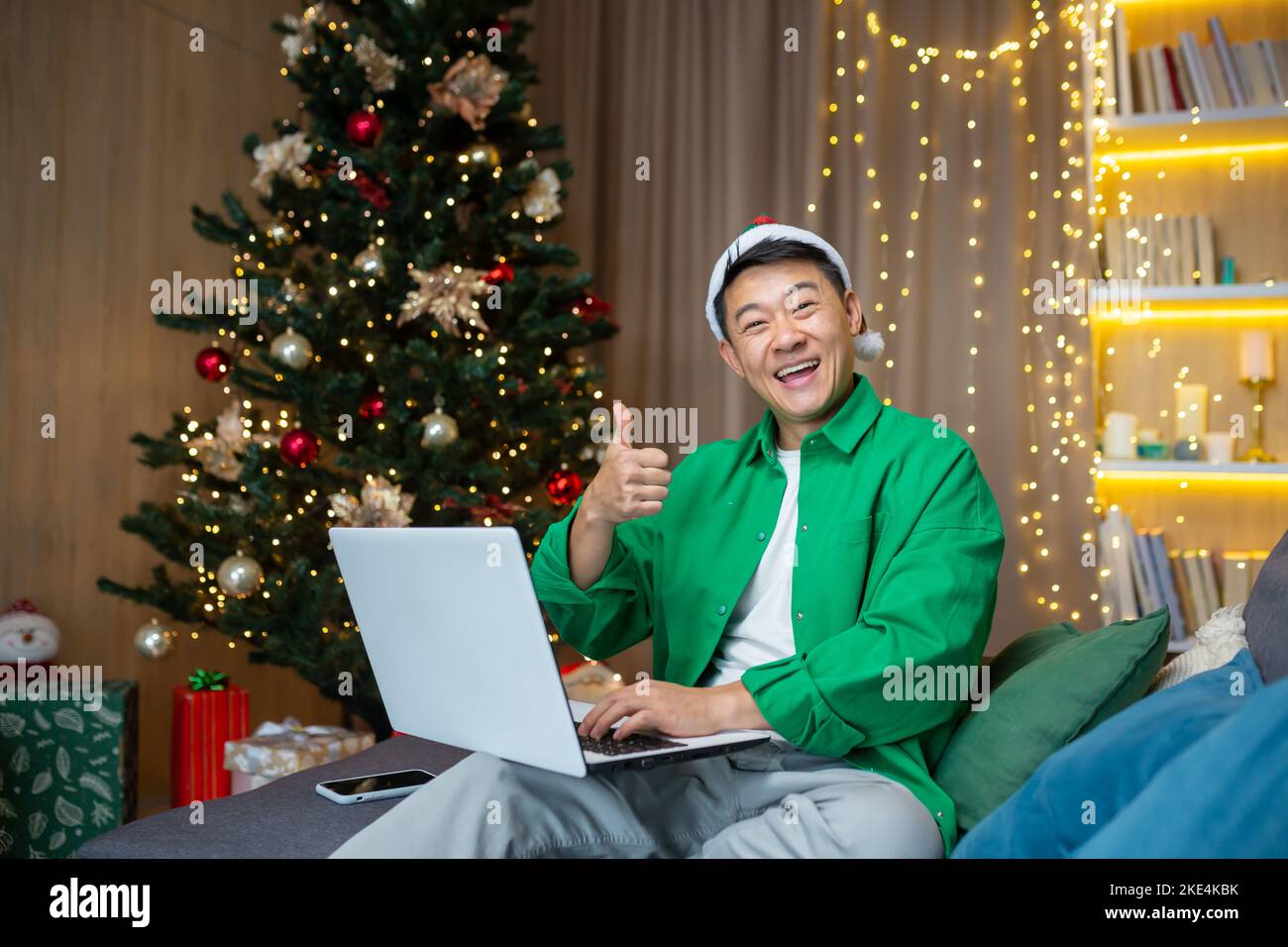Porträt eines jungen, hübschen asiatischen Mannes, der zu Hause in der Nähe des Weihnachtsbaums in einem roten Hut auf dem Sofa sitzt, einen Laptop und ein Telefon in der Hand hält, lächelnd auf die Kamera blickt und super zeigt. Stockfoto