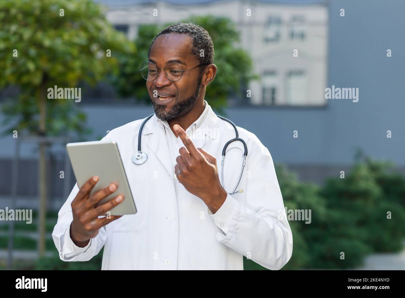Lächelnder, reifer Arzt in einem medizinischen Mantel und einer Brille mit Tablet, der per Videoanruf online per Fernzugriff spricht, afroamerikanischer Mann außerhalb der Klinik, der Patienten konsultiert. Stockfoto