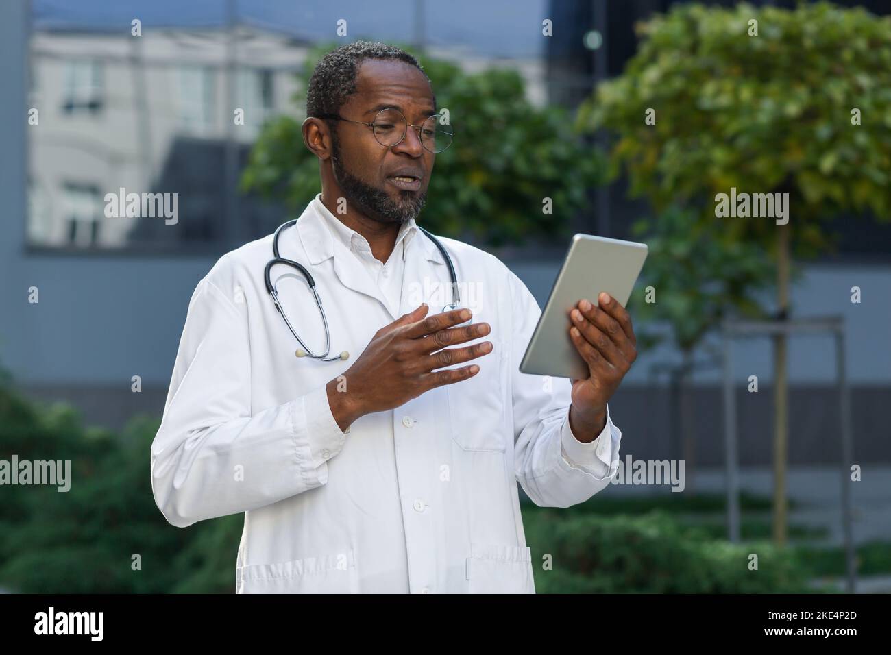 Lächelnder, reifer Arzt in einem medizinischen Mantel und einer Brille mit Tablet, der per Videoanruf online per Fernzugriff spricht, afroamerikanischer Mann außerhalb der Klinik, der Patienten konsultiert. Stockfoto