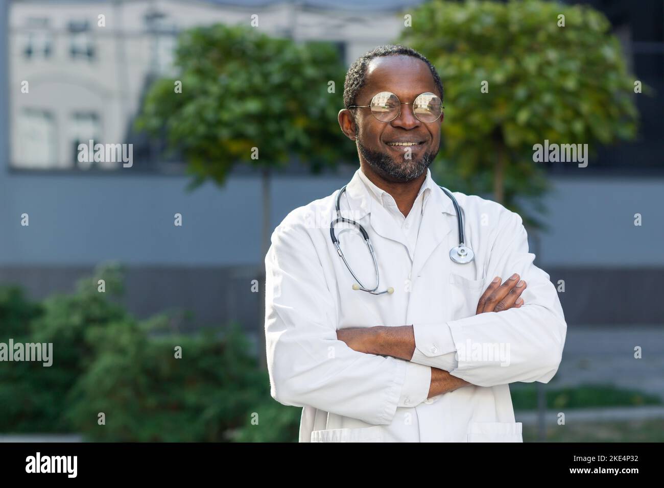 Porträt eines reifen Seniorarztes vor einer modernen Klinik blickt ein afroamerikanischer Mann mit Brille und medizinischem Mantel auf die Kamera und lächelt freundlich mit gekreuzten Armen. Stockfoto