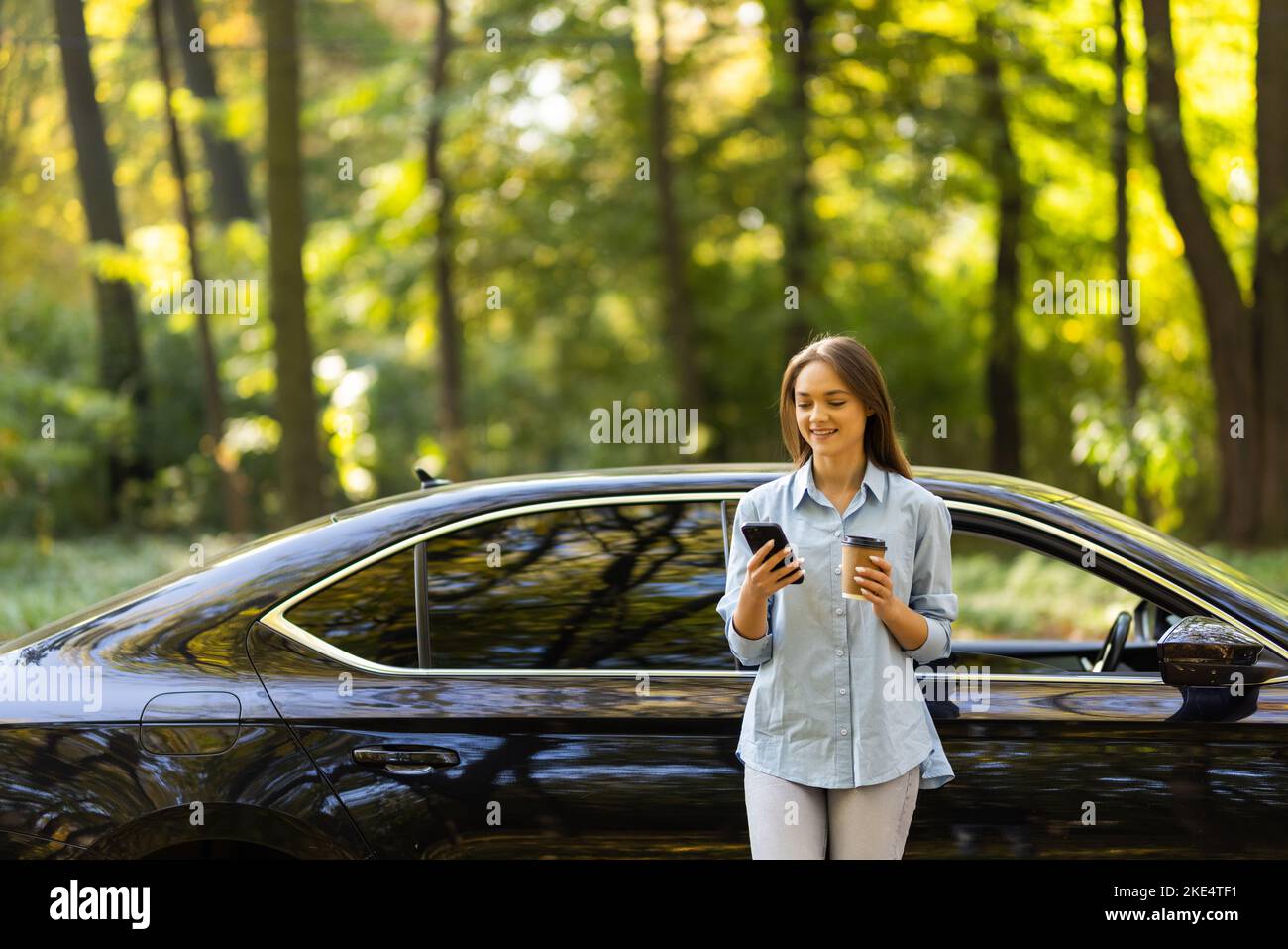 Kaukasische Frau mit einem Mobiltelefon und einer Kaffeetasse, die in der Nähe eines Elektroautos steht. Kfz-Ladung an öffentlicher Ladestation in der Nähe von Solarstrom p Stockfoto