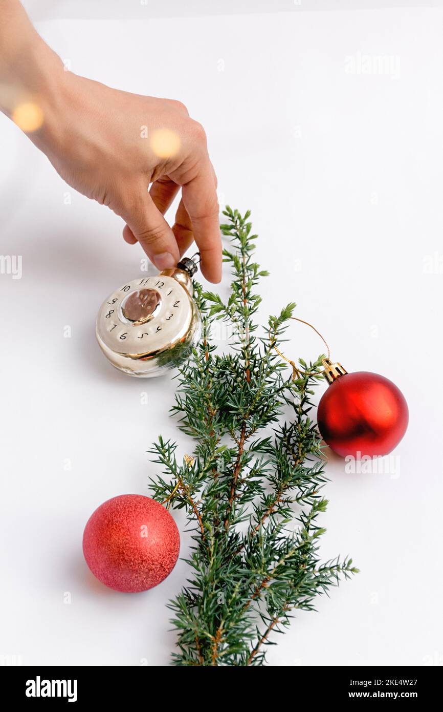 Eine weibliche Hand schmückt den Weihnachtsbaum mit roten runden Weihnachtsspielzeugen und einer Uhr auf weißem Hintergrund. Leuchtendes gelbes Bokeh. Platz für Text kopieren. Soft Selektiver Fokus. Stockfoto