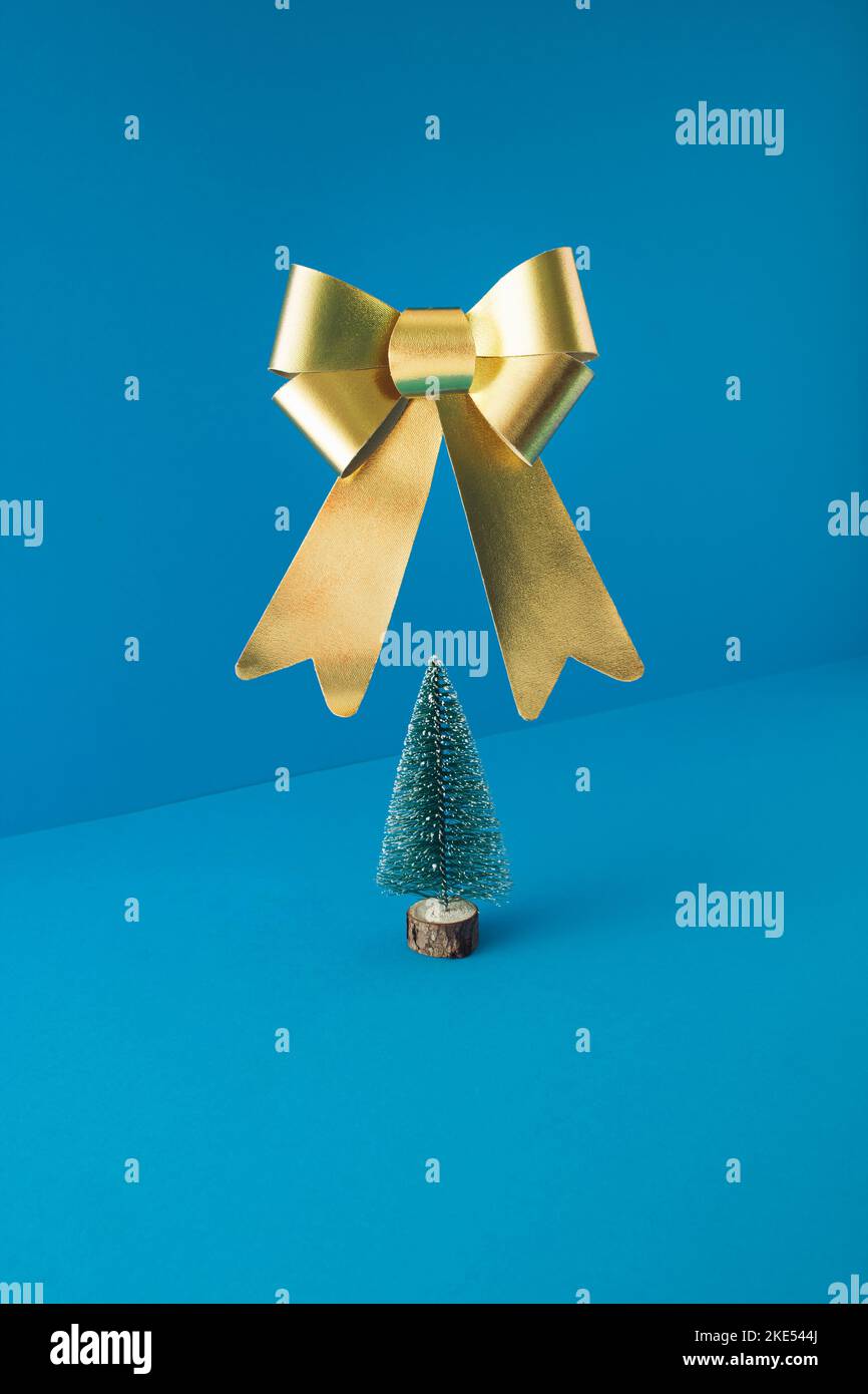 Minimale Komposition mit Weihnachtsbaum und großer goldener Schleife, die auf blauem Hintergrund schweben. Konzept für das neue Jahr. Stockfoto