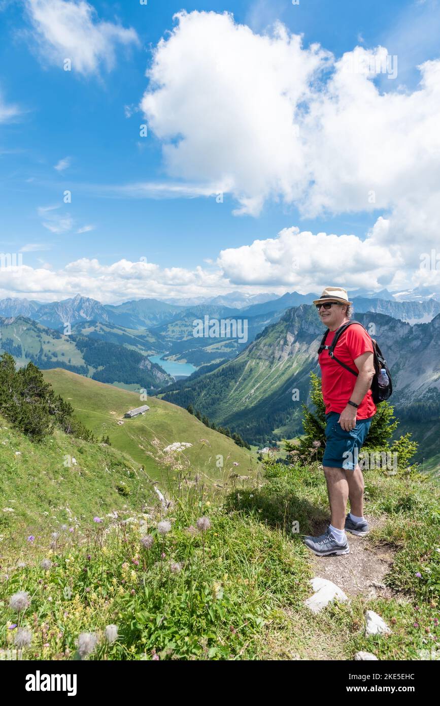 Vertikales Foto eines älteren erwachsenen Mannes mit Sonnenbrille, Mütze und Rucksack auf dem Berg mit den Schweizer Alpen Stockfoto