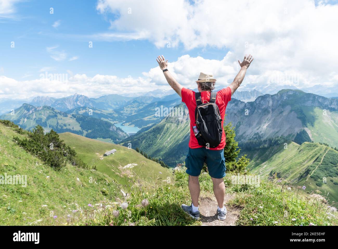 Älterer erwachsener Mann mit Hut, Rucksack und rotem Hemd, die Arme nach oben gerichtet, mit Blick auf die Schweizer Alpen. Stockfoto