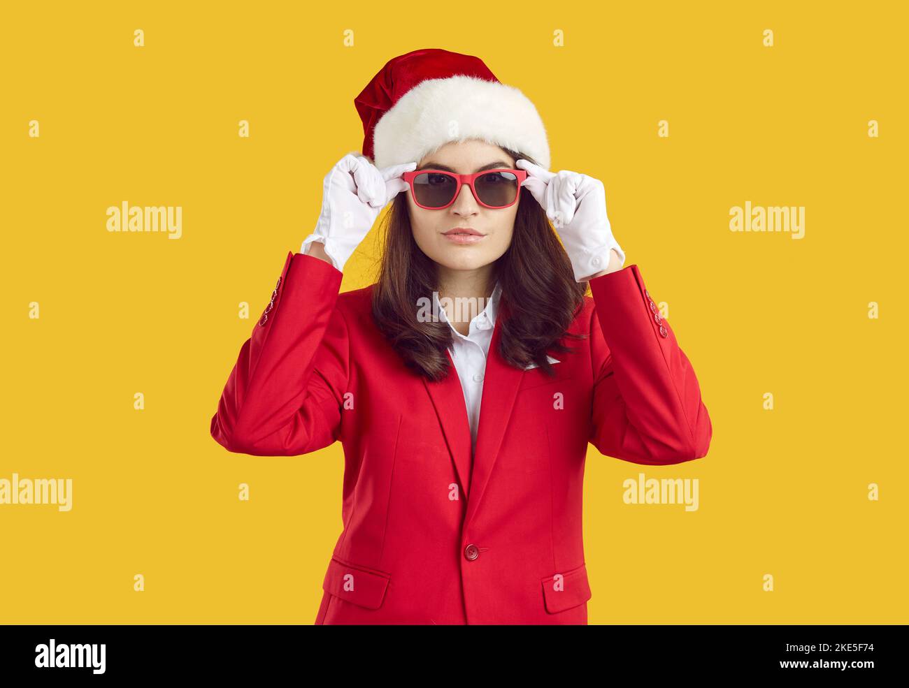 Frau in rotem Anzug, Weihnachtshut, weißen Handschuhen und Sonnenbrille isoliert auf gelbem Hintergrund Stockfoto