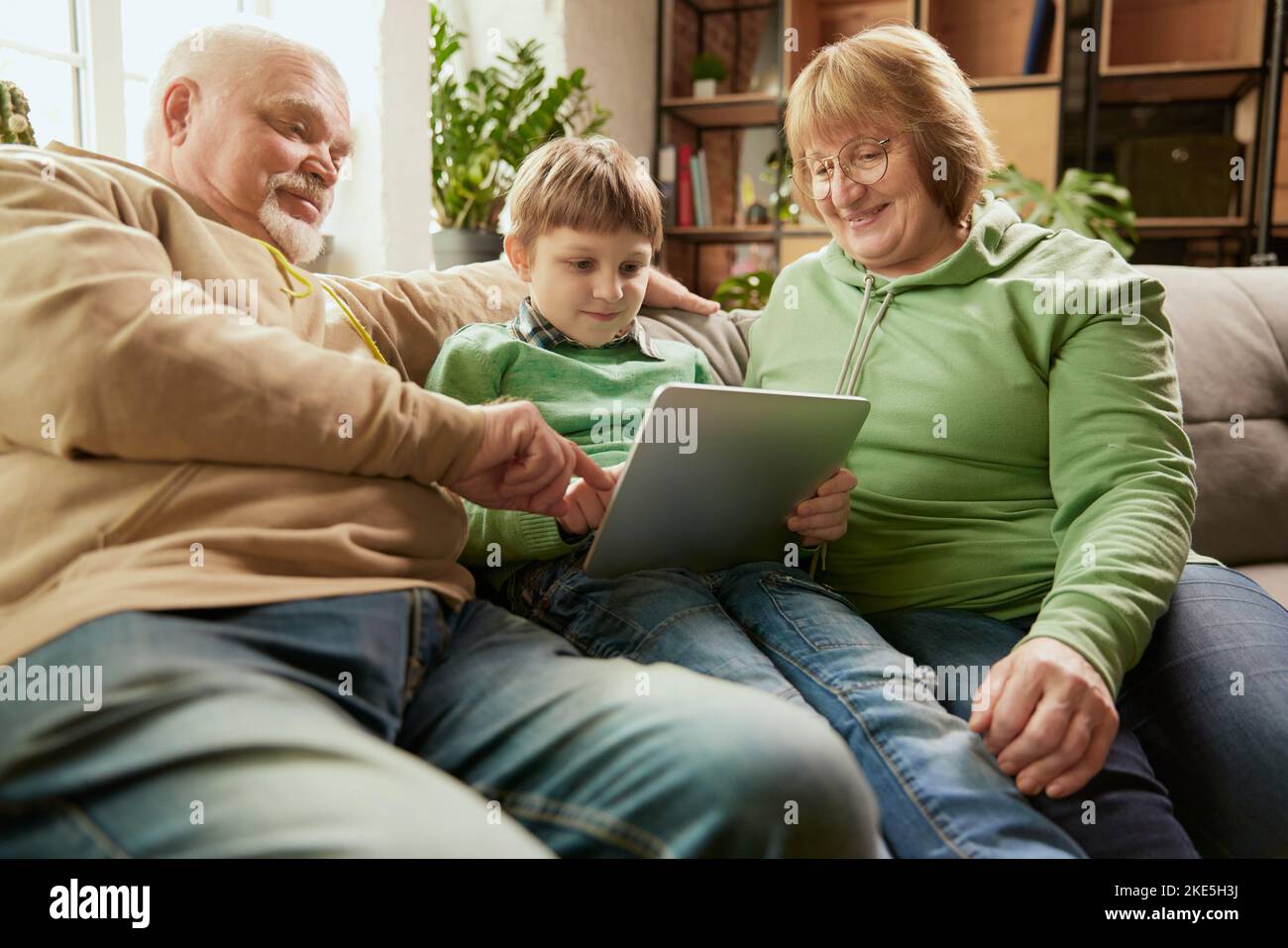 Glückliche, freundliche Familie, Großeltern und Enkel sitzen auf dem Sofa und verbringen Zeit miteinander, mit modernen Geräten, reden, studieren Stockfoto