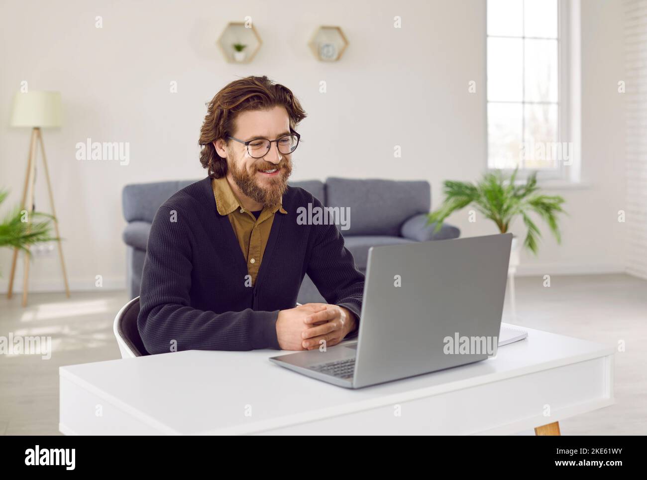 Ein positiver Mann, der mit einem Lächeln auf den Lippen auf den Laptop schaut, sitzt im Heimbüro am Schreibtisch Stockfoto