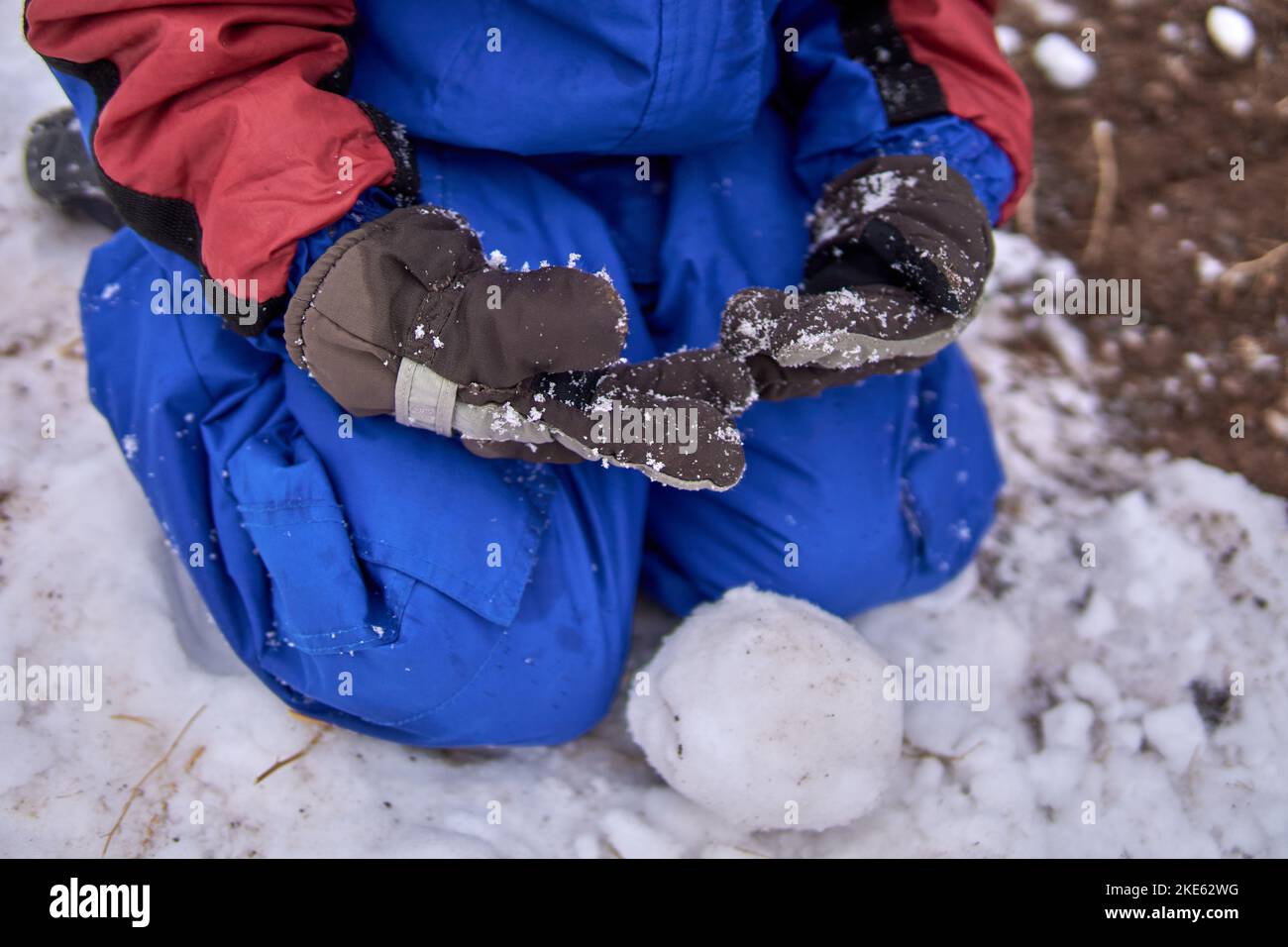 Knabe, der im Gebäude einen Schneeball kniet, bekleidet mit einem Schneeanzug und Handschuhen. Stockfoto