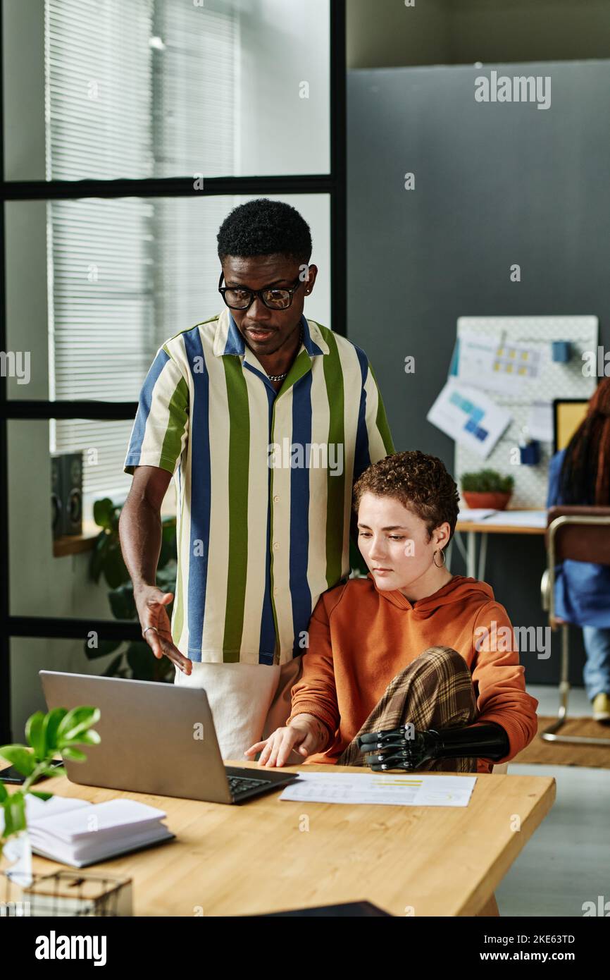 Ein junger, selbstbewusster Geschäftsmann in einem gestreiften Hemd erklärt dem Kollegen die Daten, während er während der Diskussion auf den Laptop-Bildschirm zeigt Stockfoto