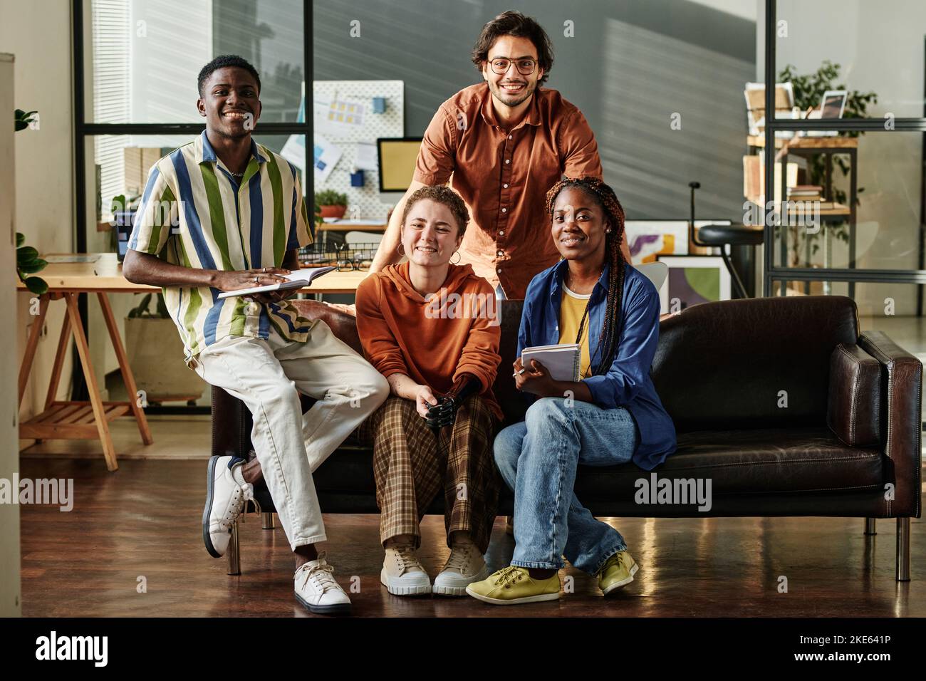 Vier junge erfolgreiche interrassische Mitarbeiter oder kreative Designer in Casualwear sitzen auf der schwarzen Ledercouch im Open-Space-Büro Stockfoto