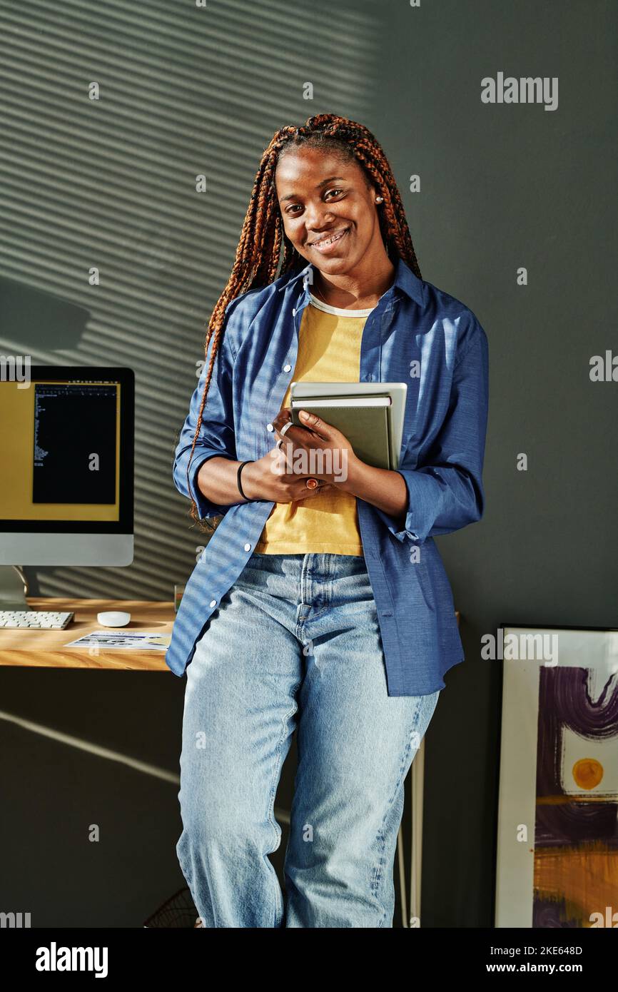 Junge afroamerikanische Geschäftsfrau in blauer Casualwear, die Notebooks hält, während sie mit einem Computermonitor am Arbeitsplatz steht Stockfoto
