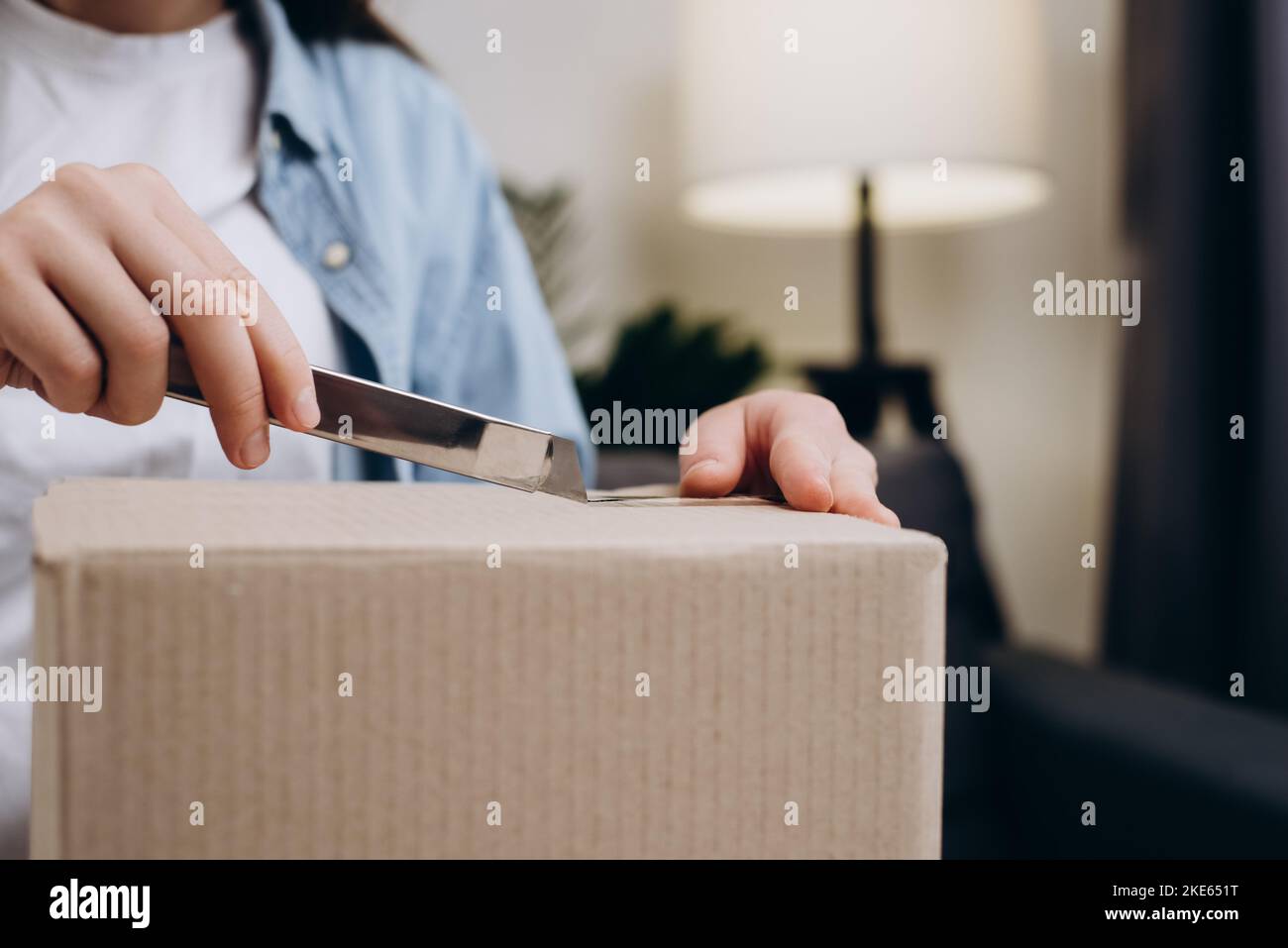 Nahaufnahme der weiblichen Hände mit dem Cutter-Messer öffnen Sie das Lieferpaket. Eine nicht wiedererkennbare junge Frau, die auf der Couch saß, erhielt eine lange erwartete Schachtel mit Geschenk. Stockfoto