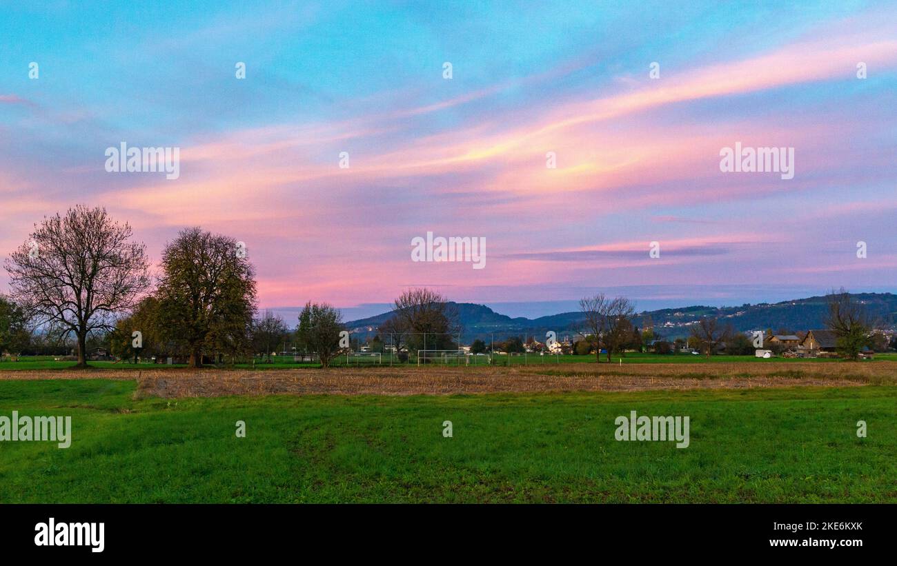 Sonnenuntergang im Rheintal, mit Wiesen und Felder, Bäumen und Schweizer Bergen im Hintergrund. Föhnwolken und blau, gelb, orange und rotem Himmel Stockfoto