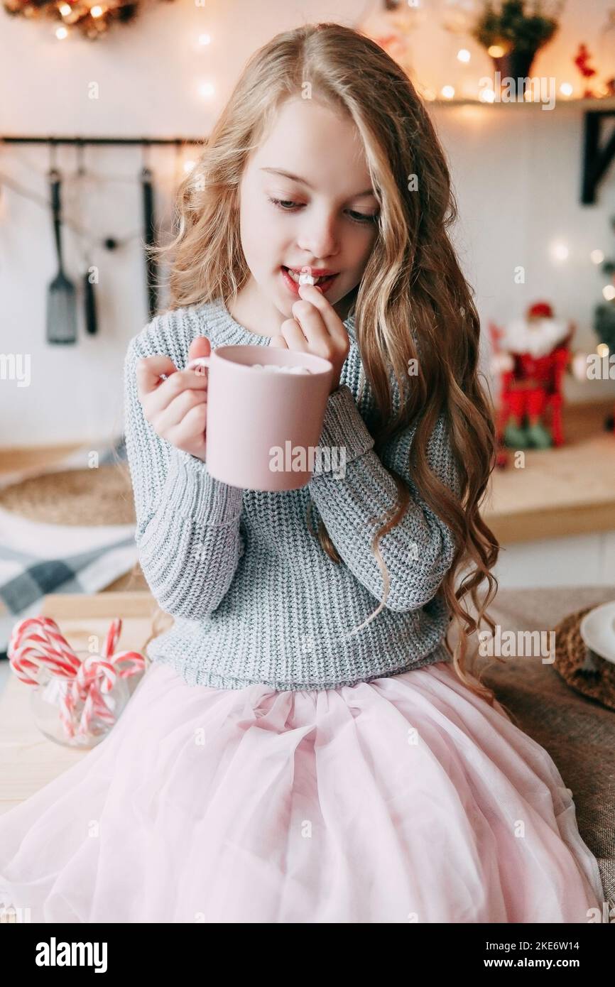 Ein blondes Mädchen mit langen Haaren trinkt mit Vergnügen Kakao aus einer rosa Tasse. Weihnachtliche Atmosphäre. Stockfoto