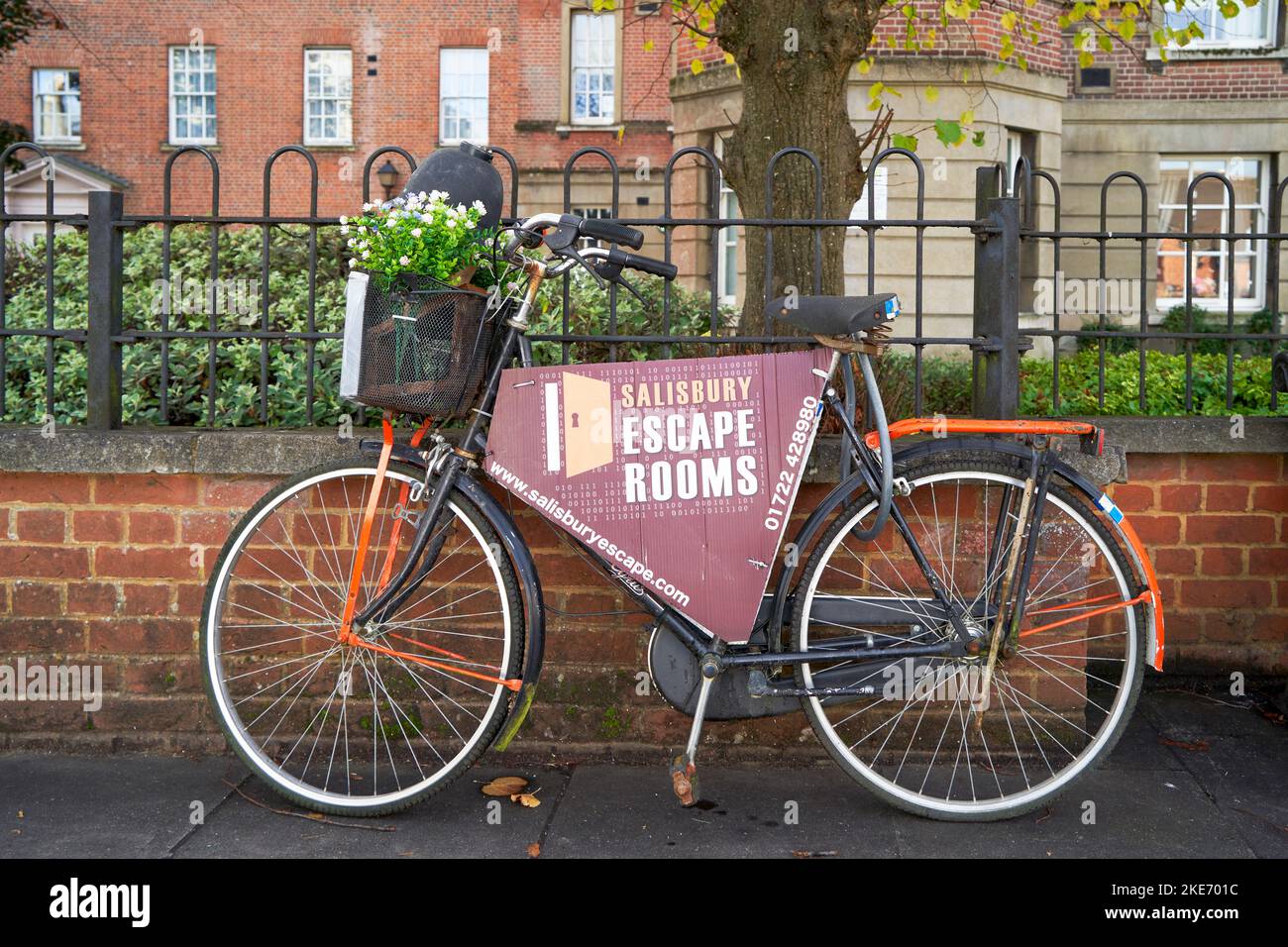 Altes Fahrrad, das an einen Metallgeländerzaun auf dem Bürgersteig gekettet wurde, der für die Werbung für ein lokales Fluchtzimmergeschäft verwendet wurde Stockfoto