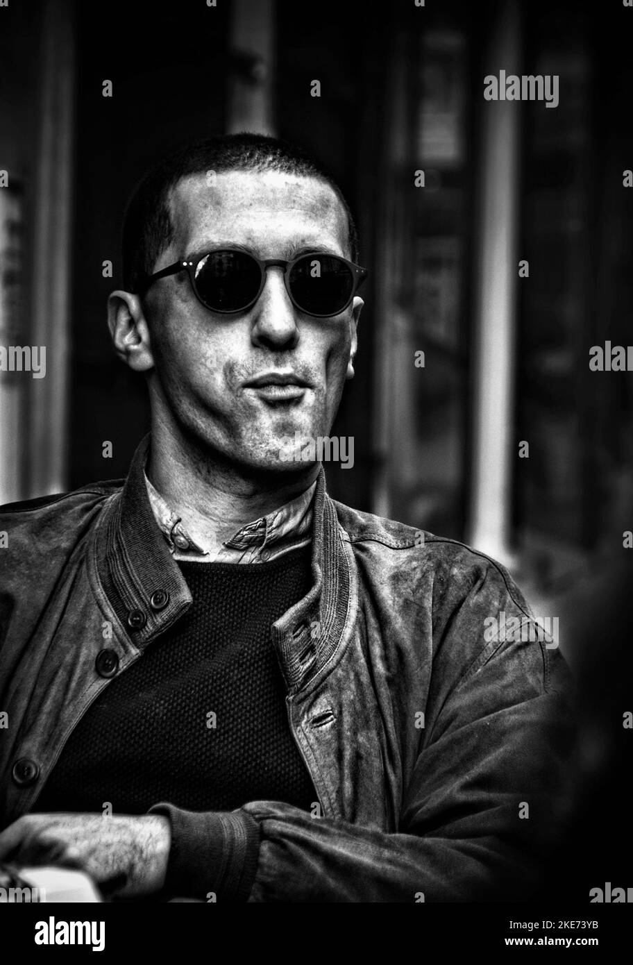 Eine vertikale Graustufe eines Mannes mit schwarzer Brille und einem Ledermantel mit verschwommenem Hintergrund Stockfoto