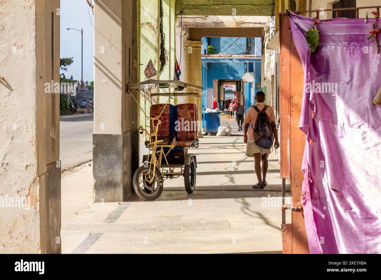 Ein violettes Tuch bedeckt die Löcher in einer metallischen Tür für Privatsphäre. Das offene bleibt unter der Veranda eines Gebäudes offen. Ein bicitaxi und Menschen sind in der Stockfoto