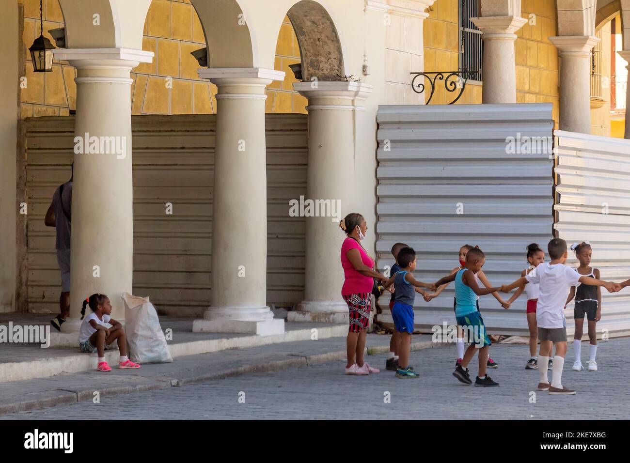 Eine Gruppe von Kindern mit einem Lehrer spielt in einer kopfsteingepflasterten Straße in Old Havanna. Ein weiteres Kind sitzt auf der Veranda eines Gebäudes. Stockfoto