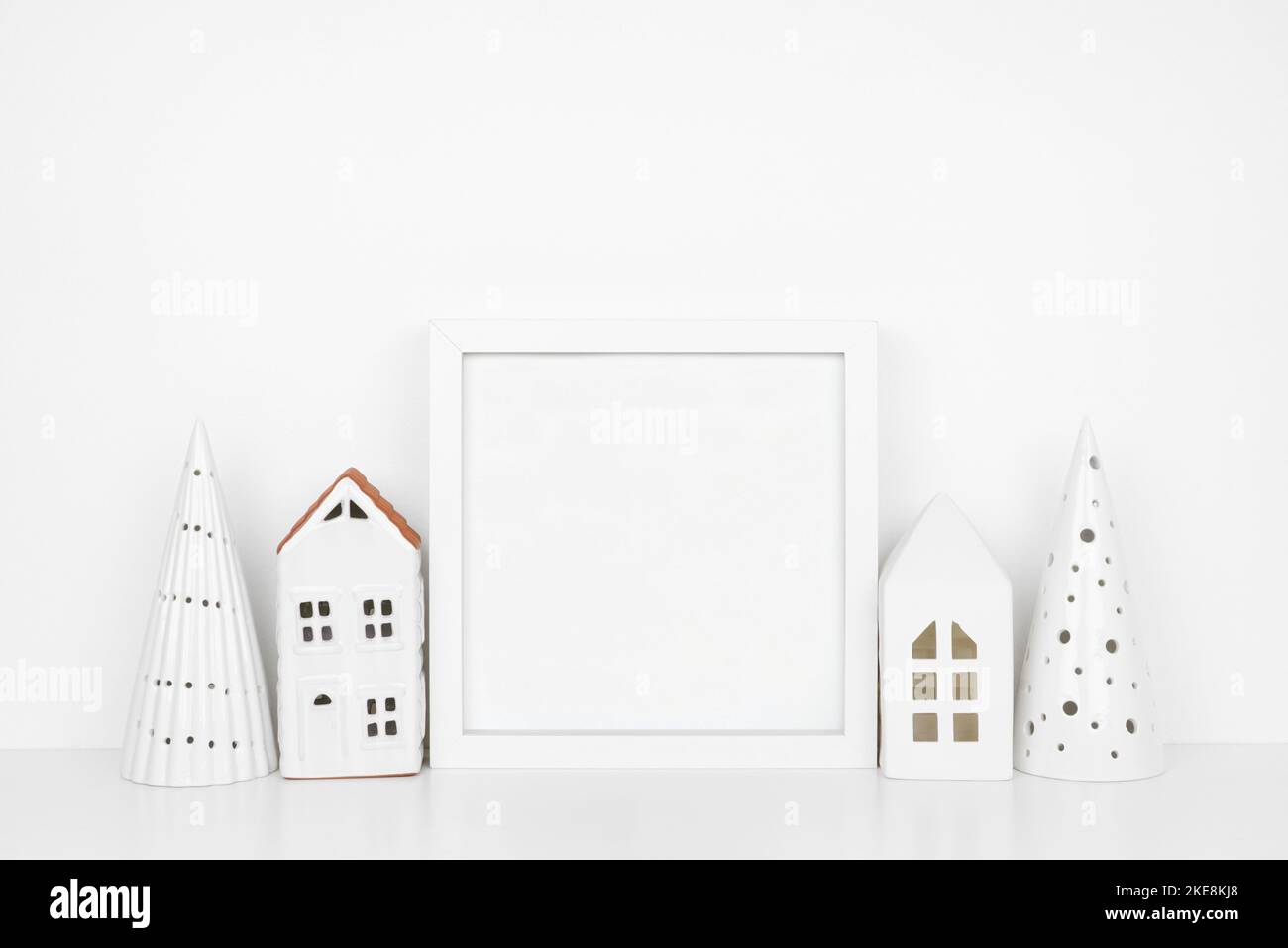 Weihnachtsmock mit weißem Rahmen und weißem Baum- und Hausdekor. Quadratischer Rahmen auf einem weißen Regal an einer weißen Wand. Speicherplatz kopieren. Stockfoto