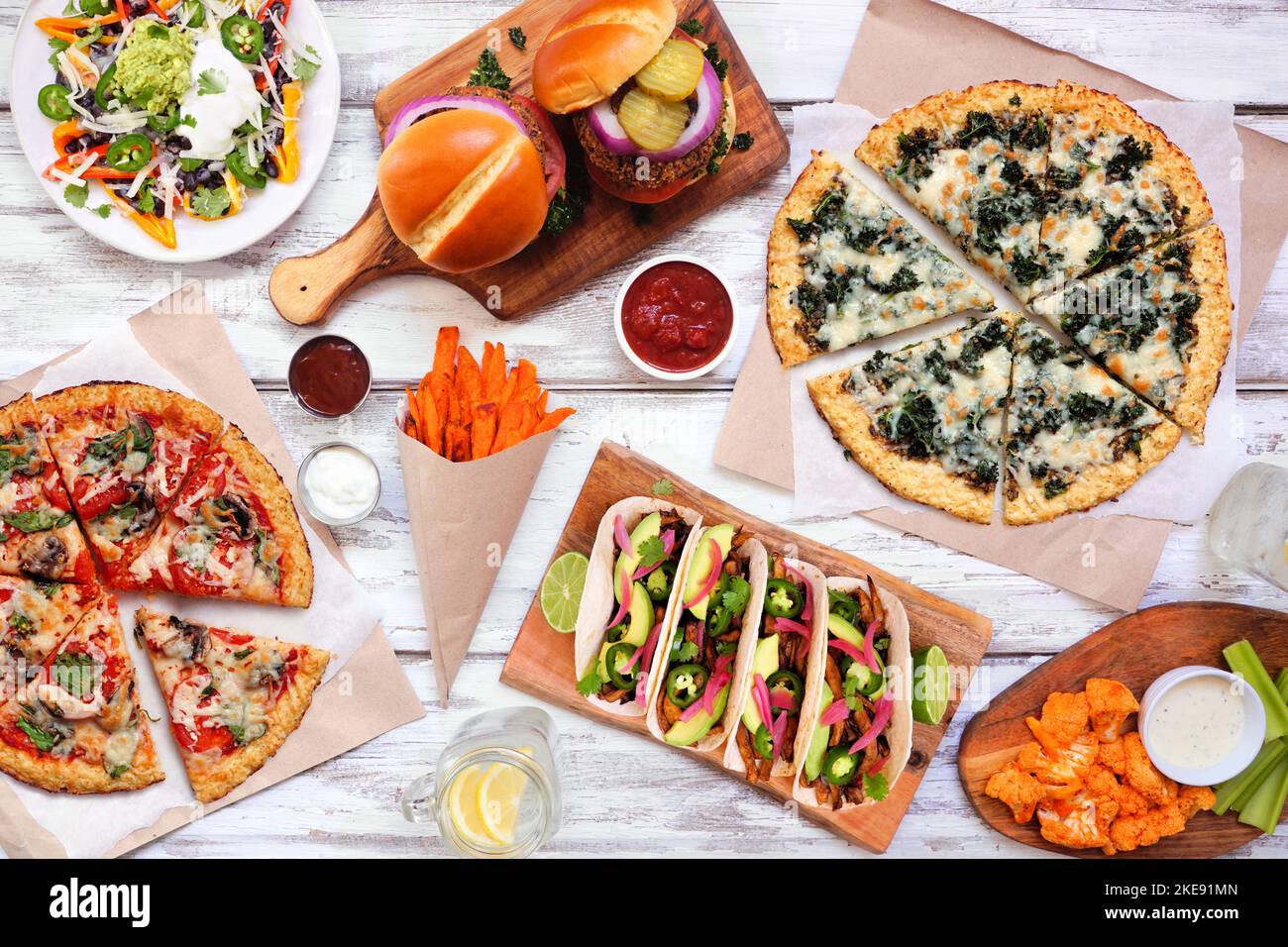 Gesunde Pflanze basiert Fast-Food-Tabelle Szene. Oben auf einem weißen Holzhintergrund. Pizzas aus Blumenkohlkruste, Bohnenburger, Pilztacos, Paprika-Nacho Stockfoto