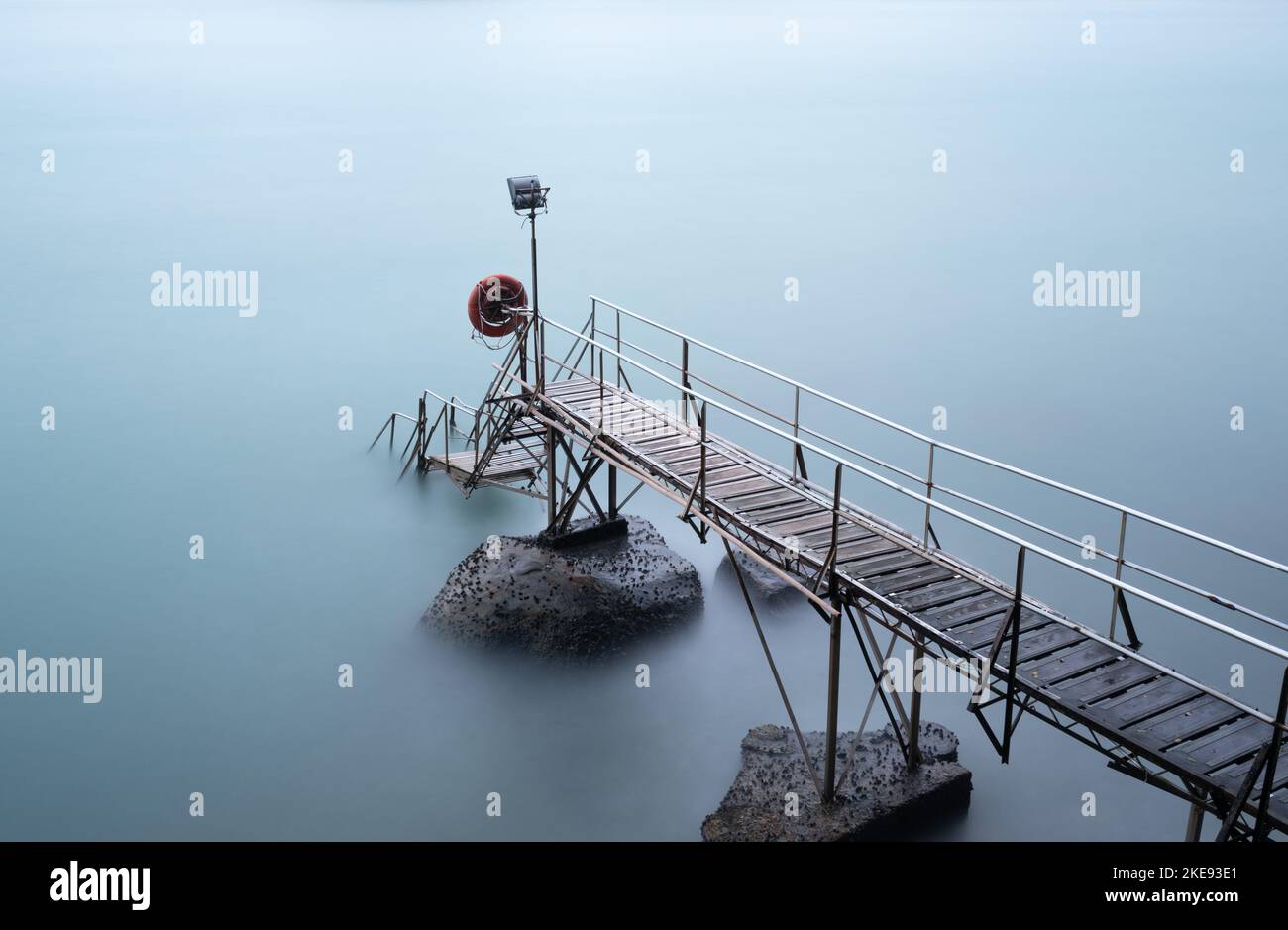 Kurzer Steg aus Metall und Holz mit Stufen, die ins Meer führen. Eine lange Belichtung verleiht dem Wasser eine seidig glatte Oberfläche Stockfoto