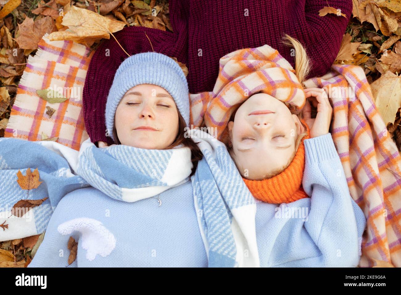 Ruhige, freundliche junge Mutter und kleine Tochter mit engen Augen in warmen blauen und orangen Outfits mit Hüten auf Laub Stockfoto