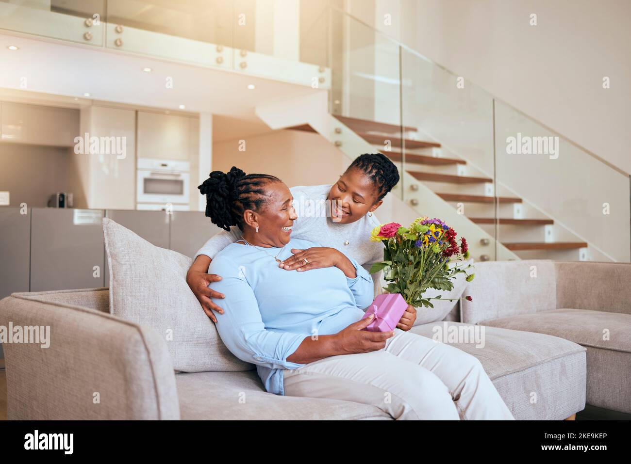 Muttertag, schwarze Frau und Überraschungsblumen auf dem Sofa als Geschenk, Schachtel oder Geschenk für Mama im Wohnzimmer. Frau, Blumenstrauß oder ältere Mama auf der Couch mit Wow Stockfoto
