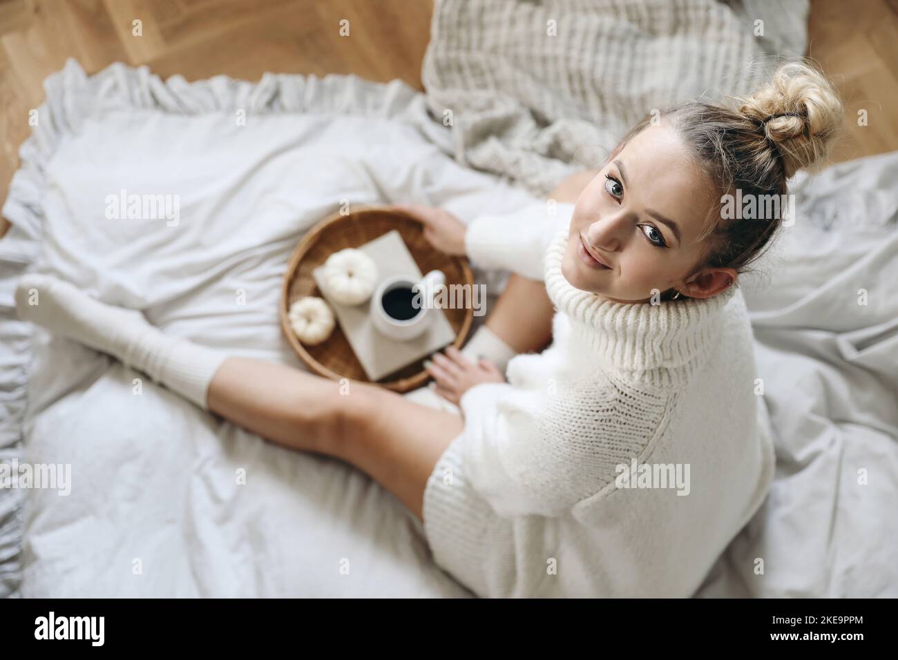 Schöne blonde kaukasische Mädchen, junge Frau in weißen Strickpullover, Socken halten Tasse Kaffee. Frühstück im Winter, Herbst im Bett. Weidenschale, weiß Stockfoto