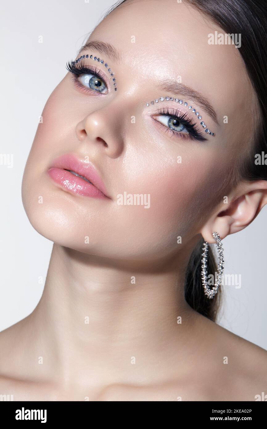 Weibliches Gesicht mit ungewöhnlichem Glitzersteinchen Make-up. Frau mit Ohrring in Form eines glänzenden Ohrrings auf grauem Hintergrund. Stockfoto