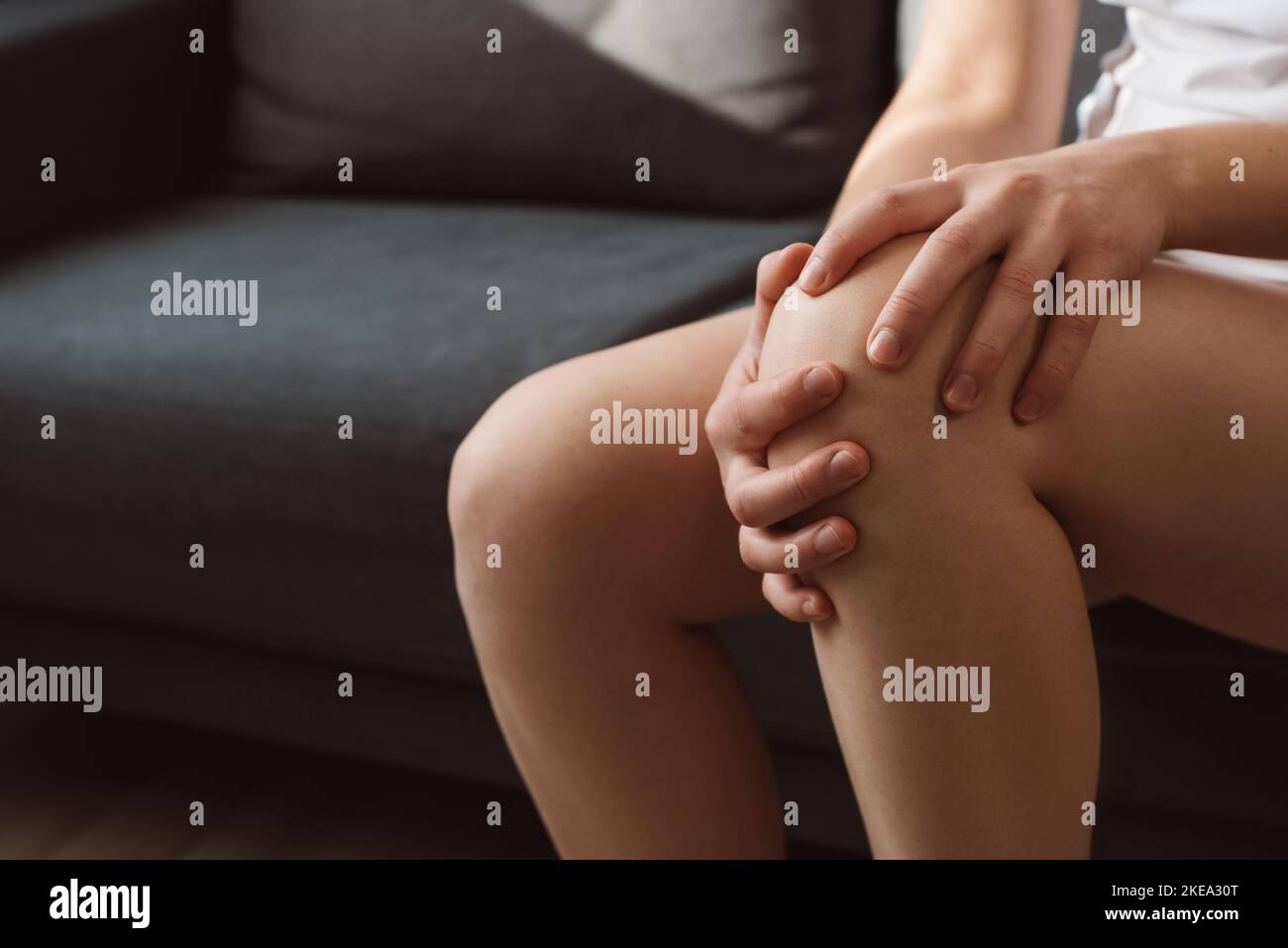 Nahaufnahme einer nicht erkennbaren jungen Frau, die zu Hause auf dem Sofa sitzt und unter Knieschmerzen leidet, massiert sich selbst. Trauma oder Bandriss. Konzept Stockfoto