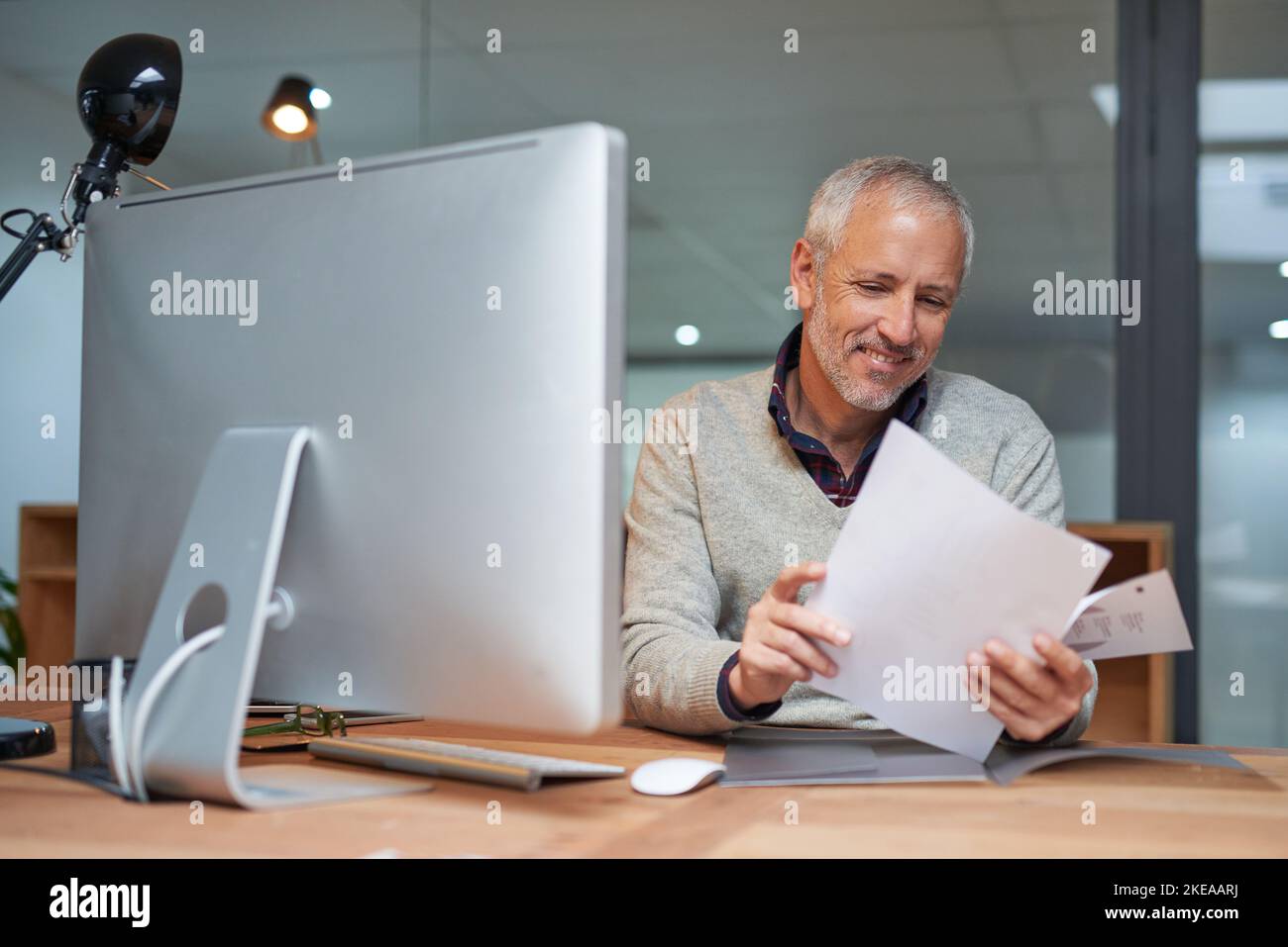Vergleich seines Arbeitsablaufs in Papierform und in digitaler Form. Porträt eines lächelnden, reifen Geschäftsmannes, der Papierkram liest, während er an seinem Schreibtisch in einem Büro sitzt. Stockfoto