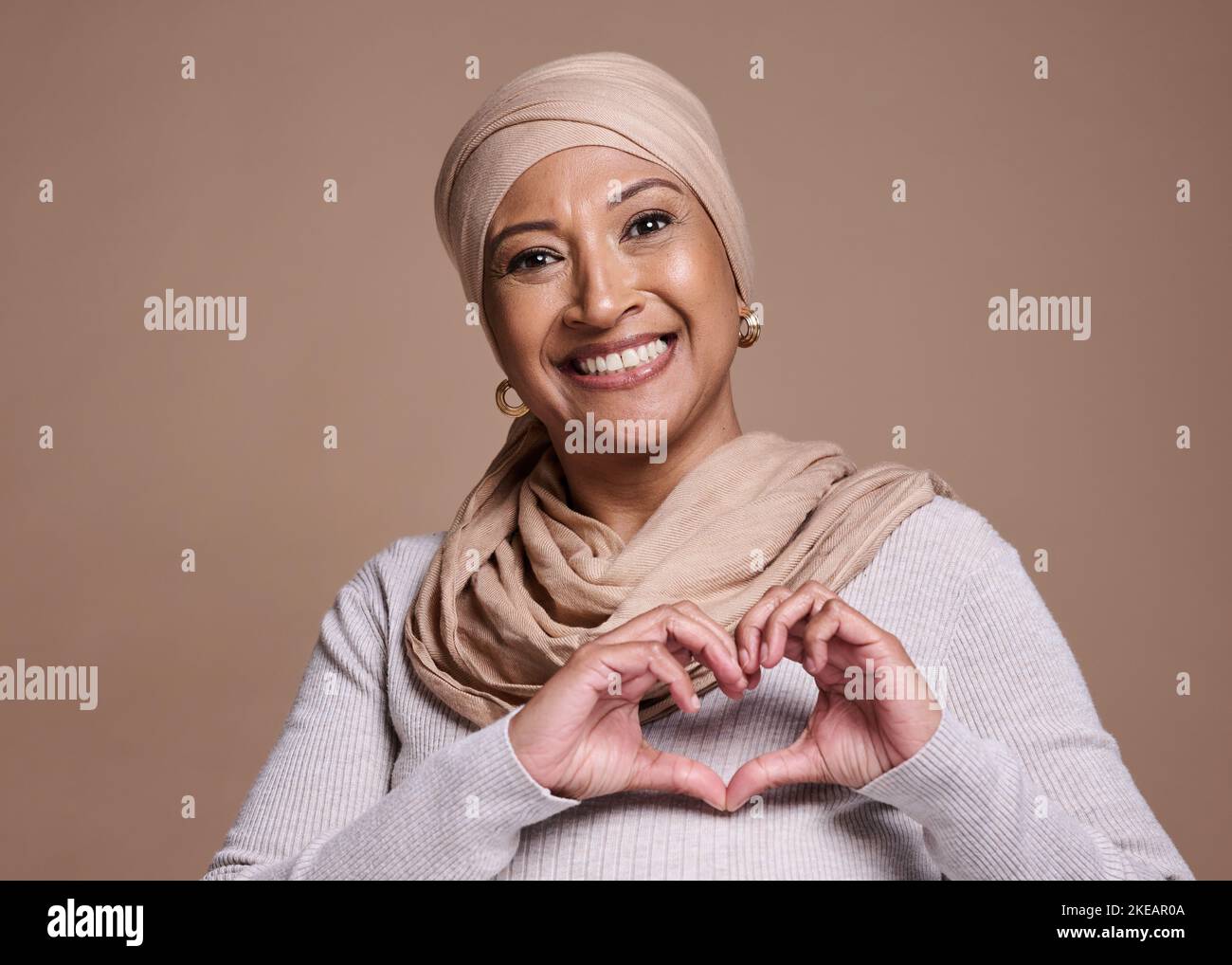 Hände, Herz und muslim mit einer reifen Frau im Studio auf braunem Hintergrund, um Liebe oder Gesundheit zu fördern. Porträt, Handzeichen und Islam mit einem Senior Stockfoto