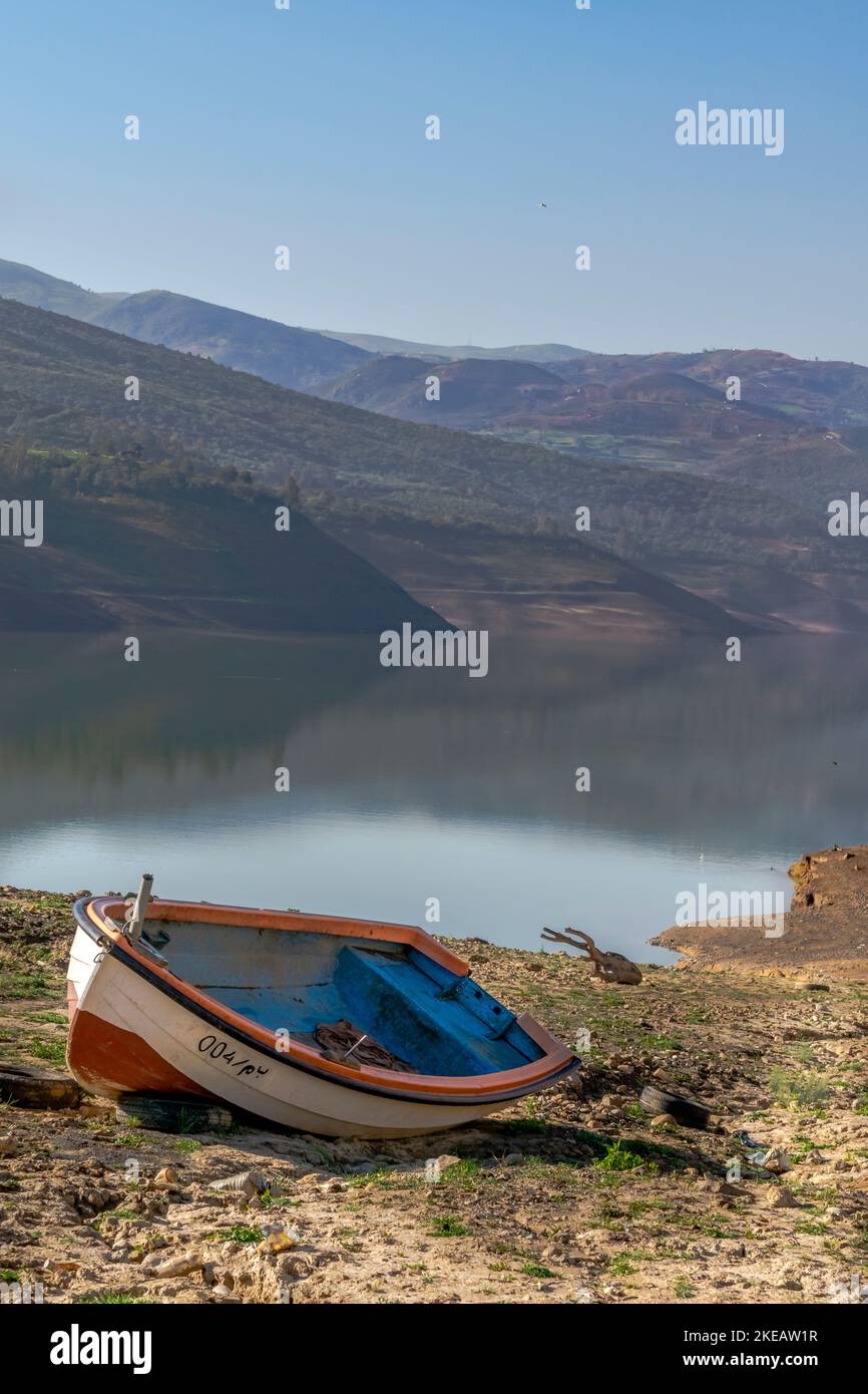 Altes hölzernes Kanu, blaues, orangefarbenes und weißes Boot, das am trockenen Ufer des Bouzegza Keddara-Staudamms gestrandet ist und neblige Berge reflektiert. Stockfoto