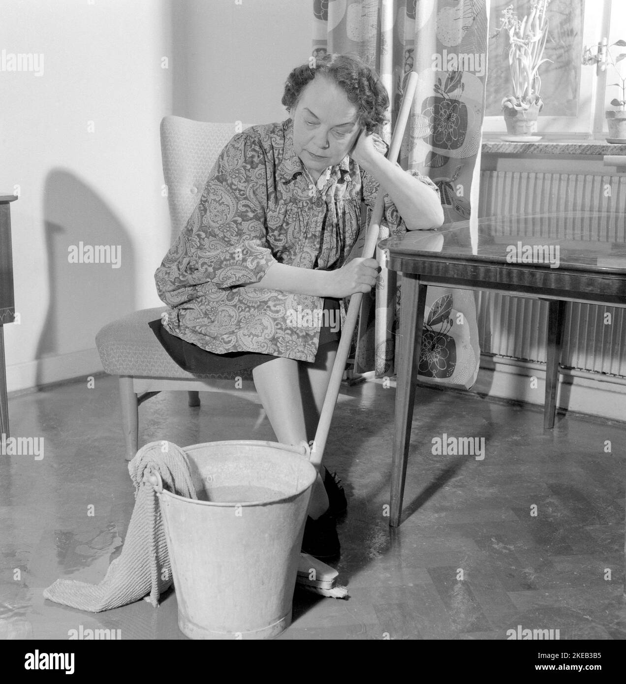 Reinigen des Bodens damals. Eine Frau sieht sehr müde und abgenutzt aus, wenn sie auf einem Stuhl sitzt und sich bei der Reinigung des Fußbodens ausruht. Schweden 1956 Conard Ref. 3160 Stockfoto