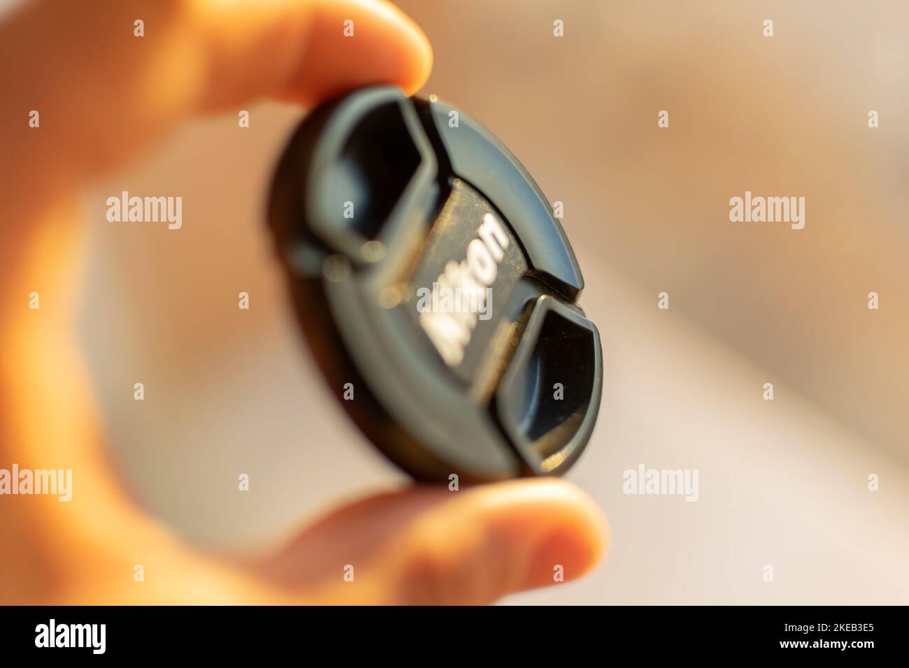 Makrofotografie einer Nikon-Frontlinsenabdeckung, die zwischen den Fingern in goldenem Sonnenlicht reflektiert wird. Verschwommener Vorder- und Hintergrund Stockfoto