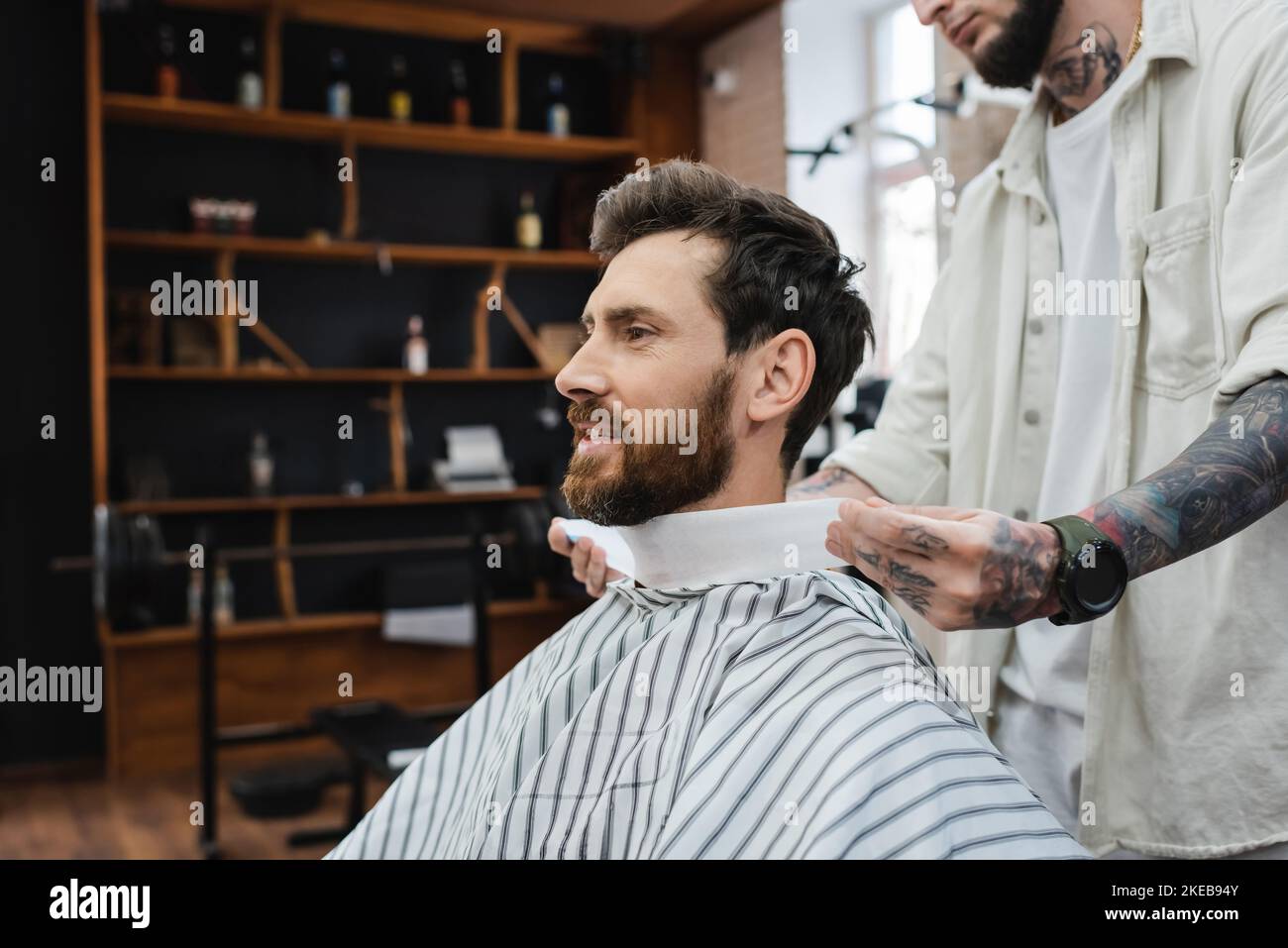 Tätowierte Barbier, der den Hals des bärtigen Mannes mit Haarreif umwickelt, Stockbild Stockfoto