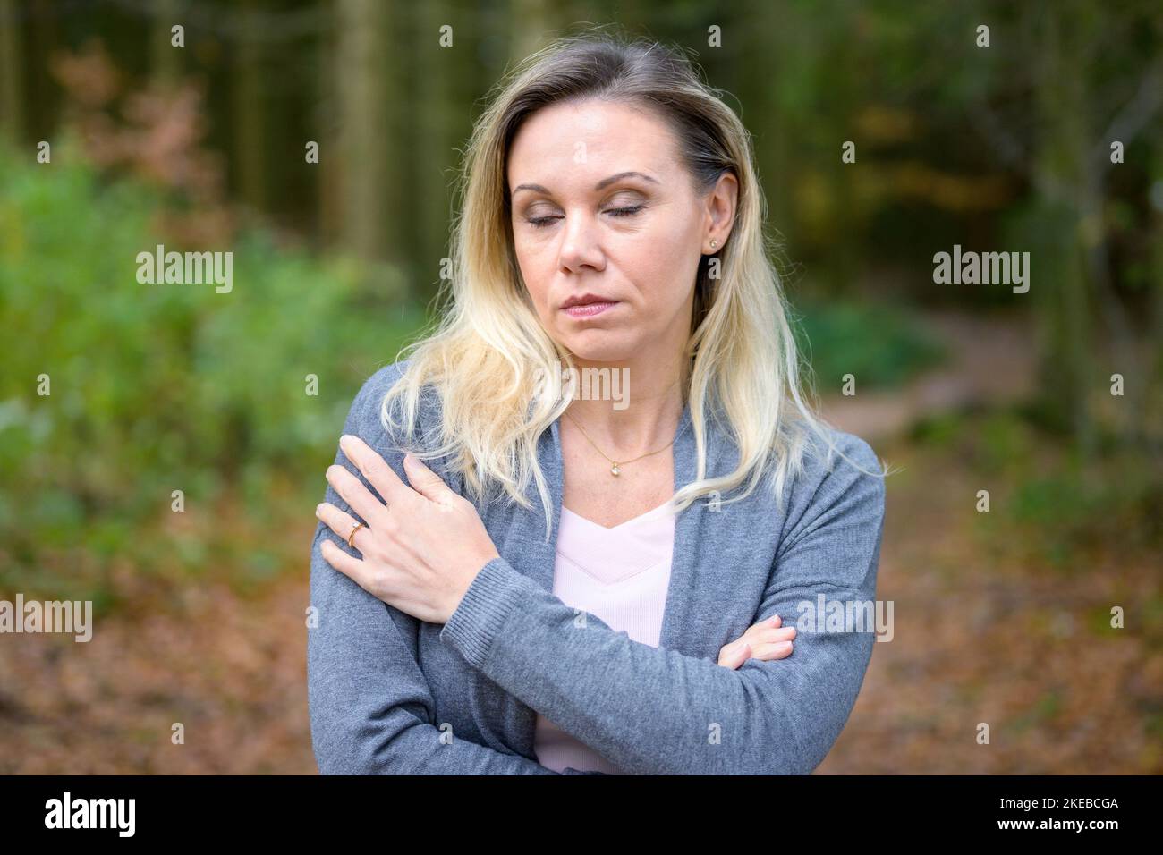 Schöne blonde Frau mittleren Alters, die in einem Wald mit gekreuzten Armen und geschlossenen Augen steht Stockfoto