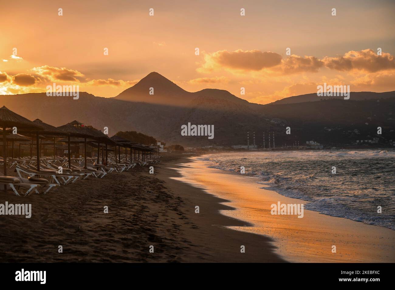 Amoudara Strand in Sonnenuntergang Beleuchtung. Sandstrand, Sonnenschirmreihen, blauwelliges Meerwasser mit orangefarbener Sonnenreflexion, Berge mit dem höchsten Berg Strou Stockfoto