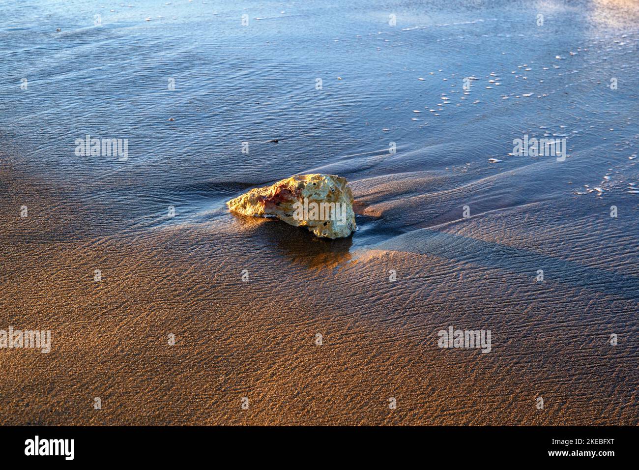 Nasser kleiner Stein auf nassem Strandsand, viele schlangen im Sand von Wasser und Wind. In blauer, gelber und brauner Farbe. Stockfoto
