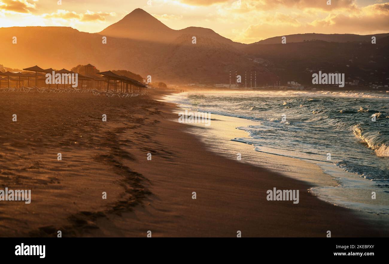 Moody Sonnenuntergang am Strand. Sand, Sonnenschirmreihen, blauwelliges Meerwasser, Berge in schöner Beleuchtung, höchster Hügel Stroumpoulas. Amoudara Beach, Kreta, GRE Stockfoto