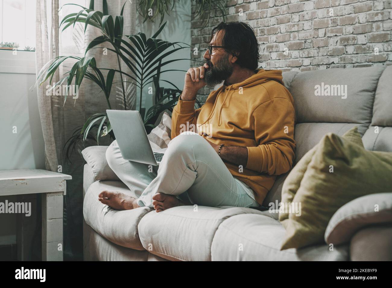 Reifer Mann, der zu Hause mit gekreuzten Beinen sitzt und am Laptop arbeitet. Moderne Menschen mit Computer auf dem Sofa in der Wohnung. Konzept der Internetverbindung und Surfen im Internet. Intelligente Arbeitstechnologie Stockfoto
